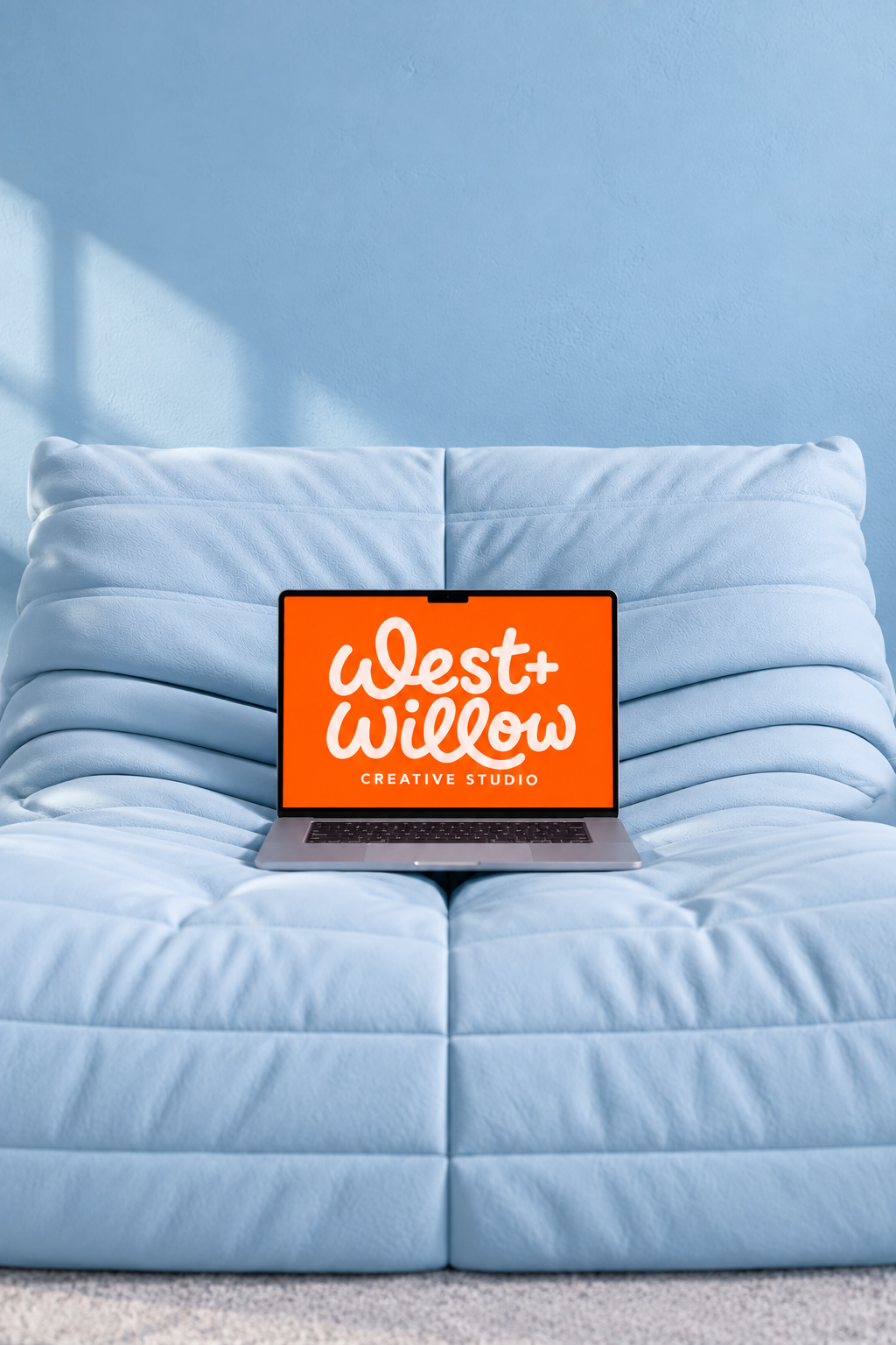 A laptop on a light blue couch with a blue wall in the background, displaying the logo of 'Welt+Willow Creative Studio' on the screen.