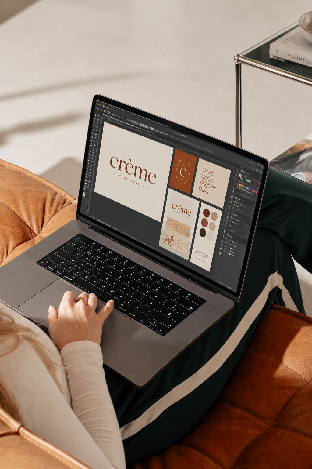 Person working on a laptop with a branding and design project for 'Crème Coffee Roasters' open on the screen, sitting on a brown leather or suede couch in a cozy space.