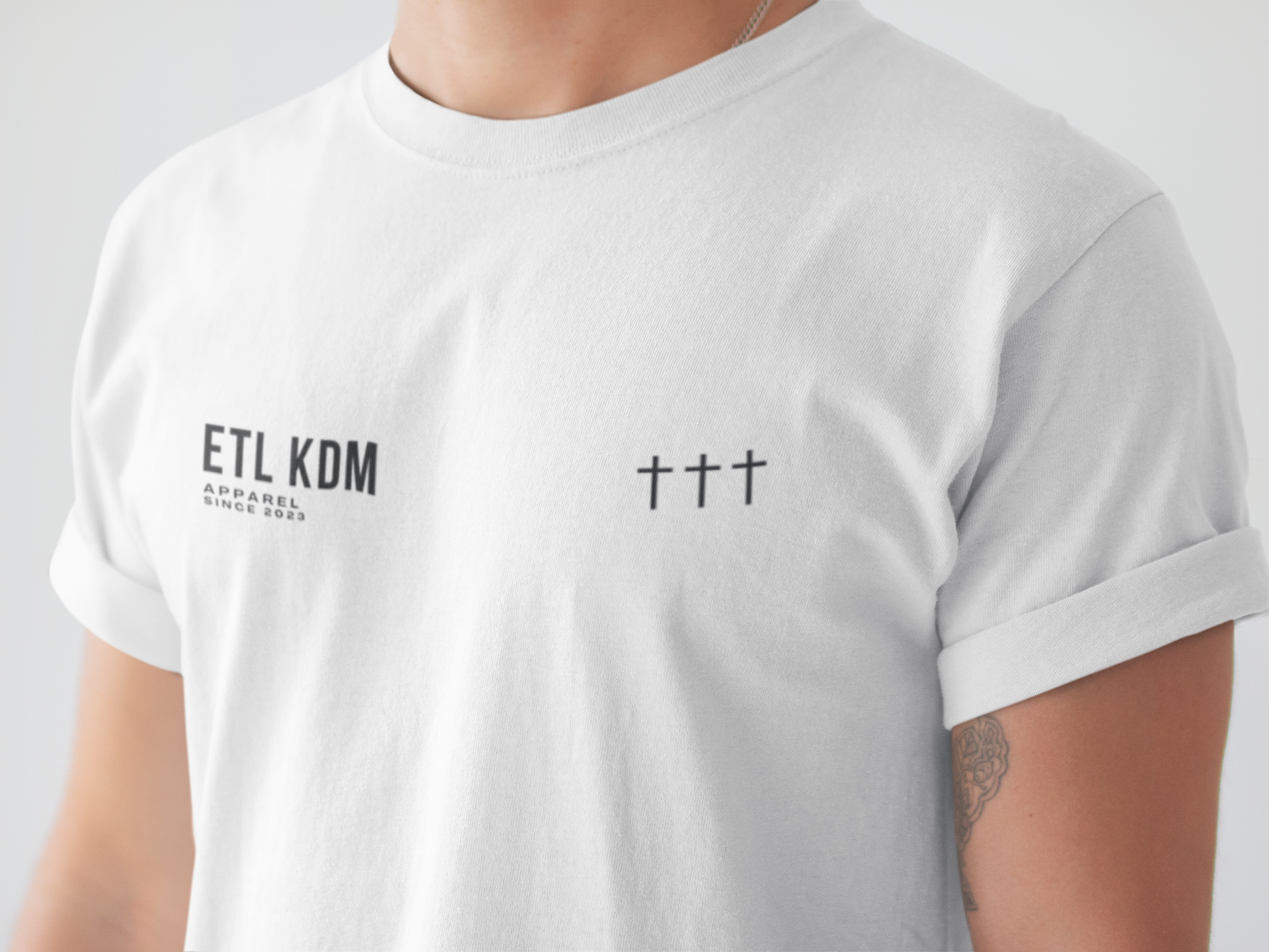 Close-up of a person wearing a white t-shirt with black text that reads 'ETL KDM APPAREL SINCE 2023,' with three black crosses on the right side of the chest, and a tattoo on the person's left arm visible near the wrist.