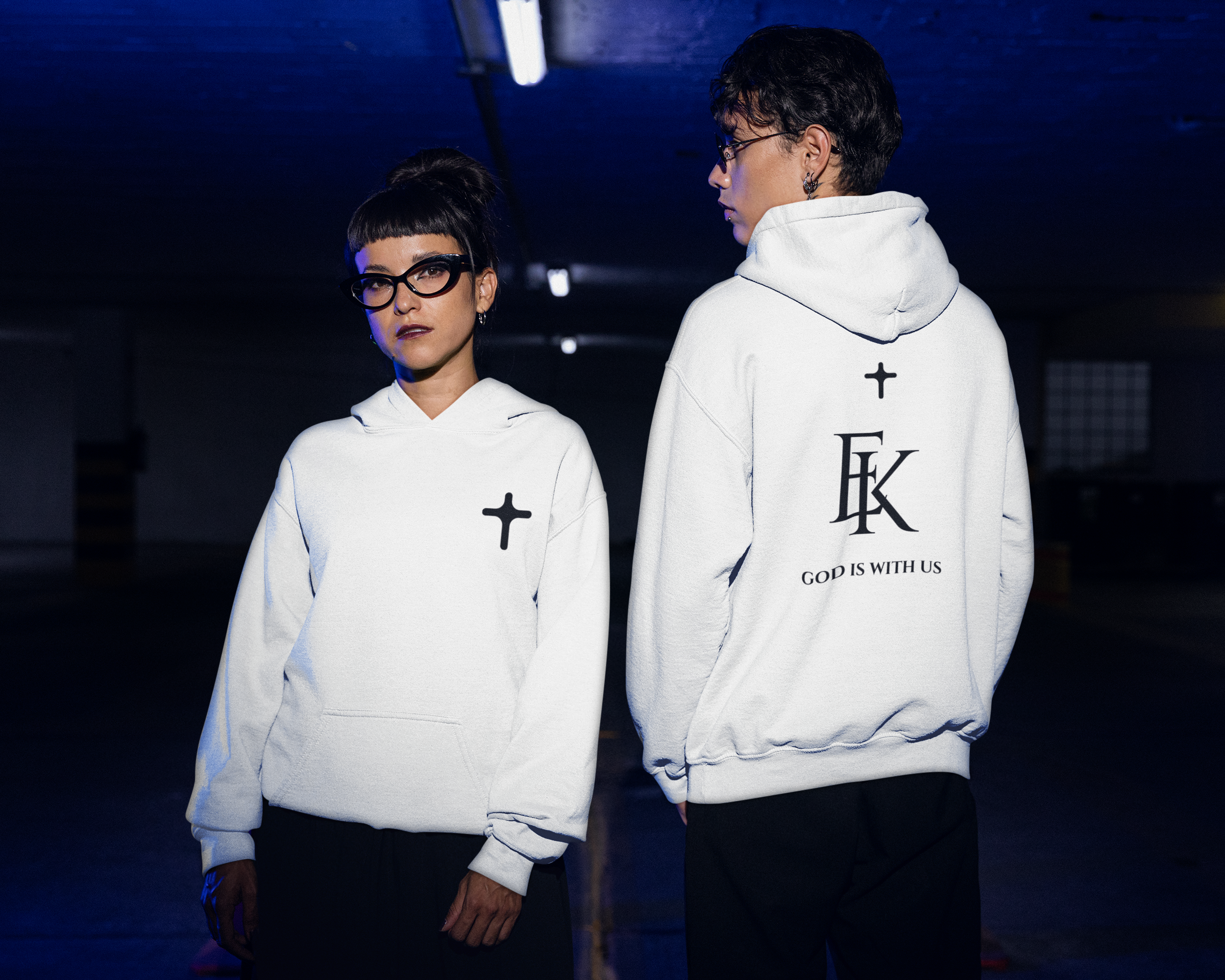 Two individuals wearing white hoodies with black text on the back, standing in a dimly lit environment with a dark blue ceiling. The woman on the left has short dark hair with bangs and is wearing black glasses, looking directly at the camera. The pe