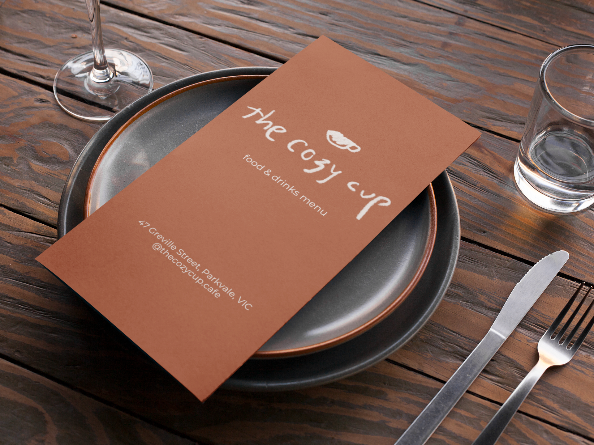 A menu titled 'The cozy cup' with a coffee cup illustration and text reading food & drinks menu, placed on a black plate on a wooden table, with a glass of water on the side and cutlery nearby.