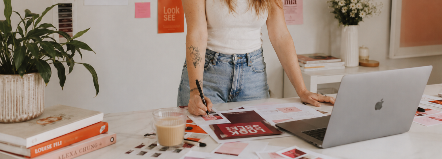Woman in white tank top and jeans standing at a cluttered white table, writing on papers with a black marker, surrounded by a MacBook, coffee, books, and a plant.