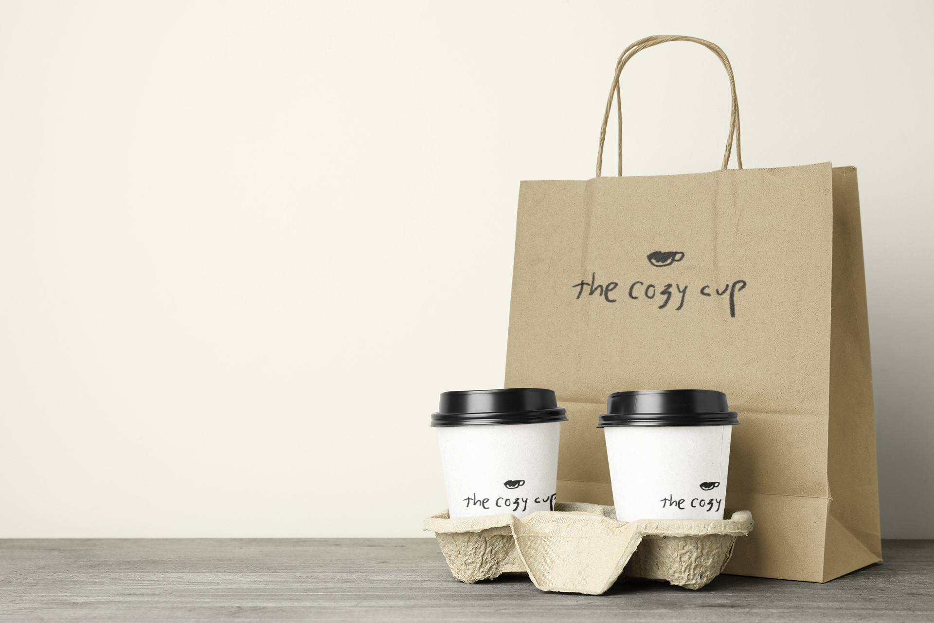 Two white coffee cups with black lids, a cardboard coffee carrier, and a paper shopping bag with the words "the cozy cup" and a coffee bean logo on it, placed on a gray wooden surface against a beige background.