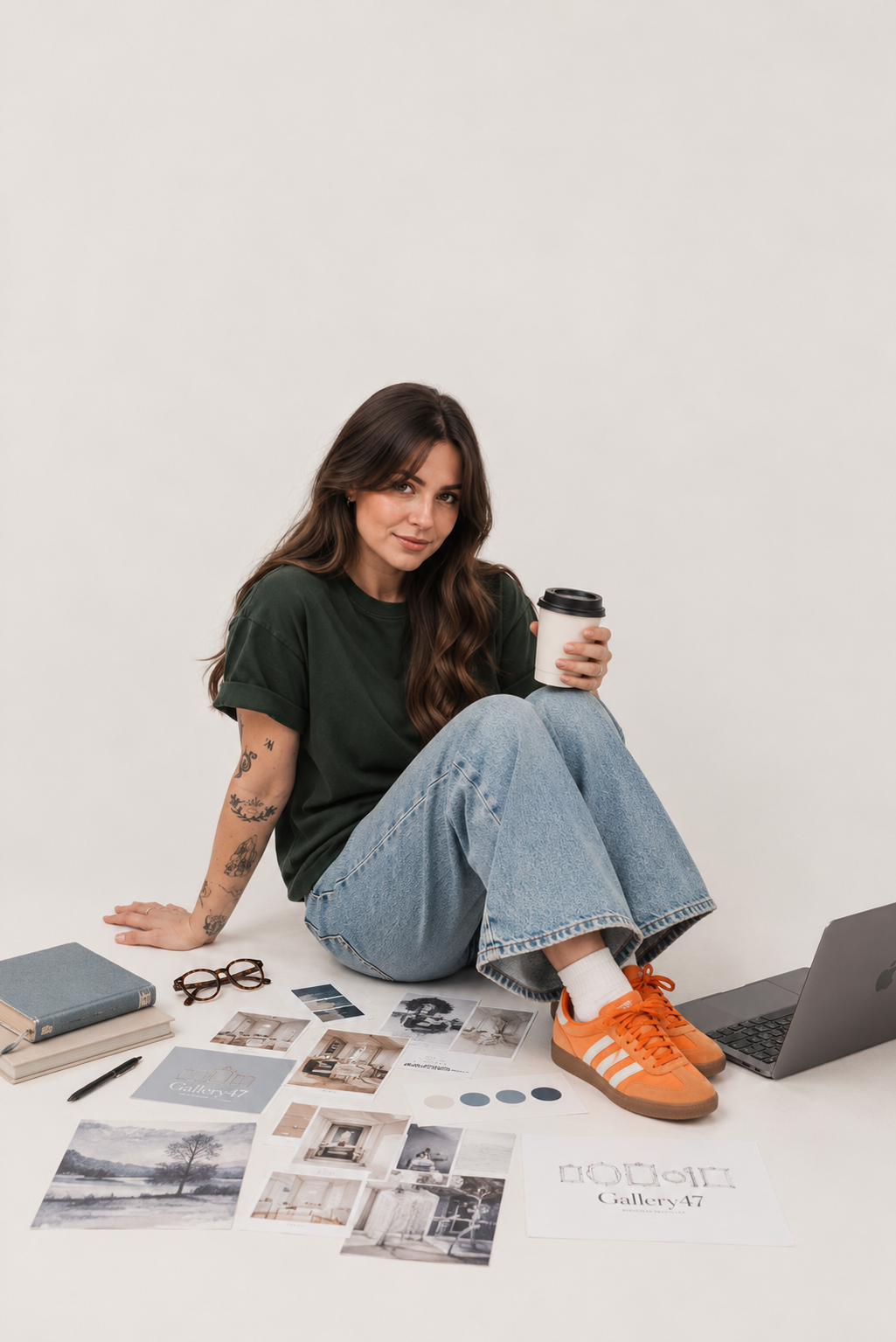 Young woman with tattoos, wearing a green t-shirt and jeans, sitting on the floor with photos, a notebook, and a laptop, holding a coffee cup.