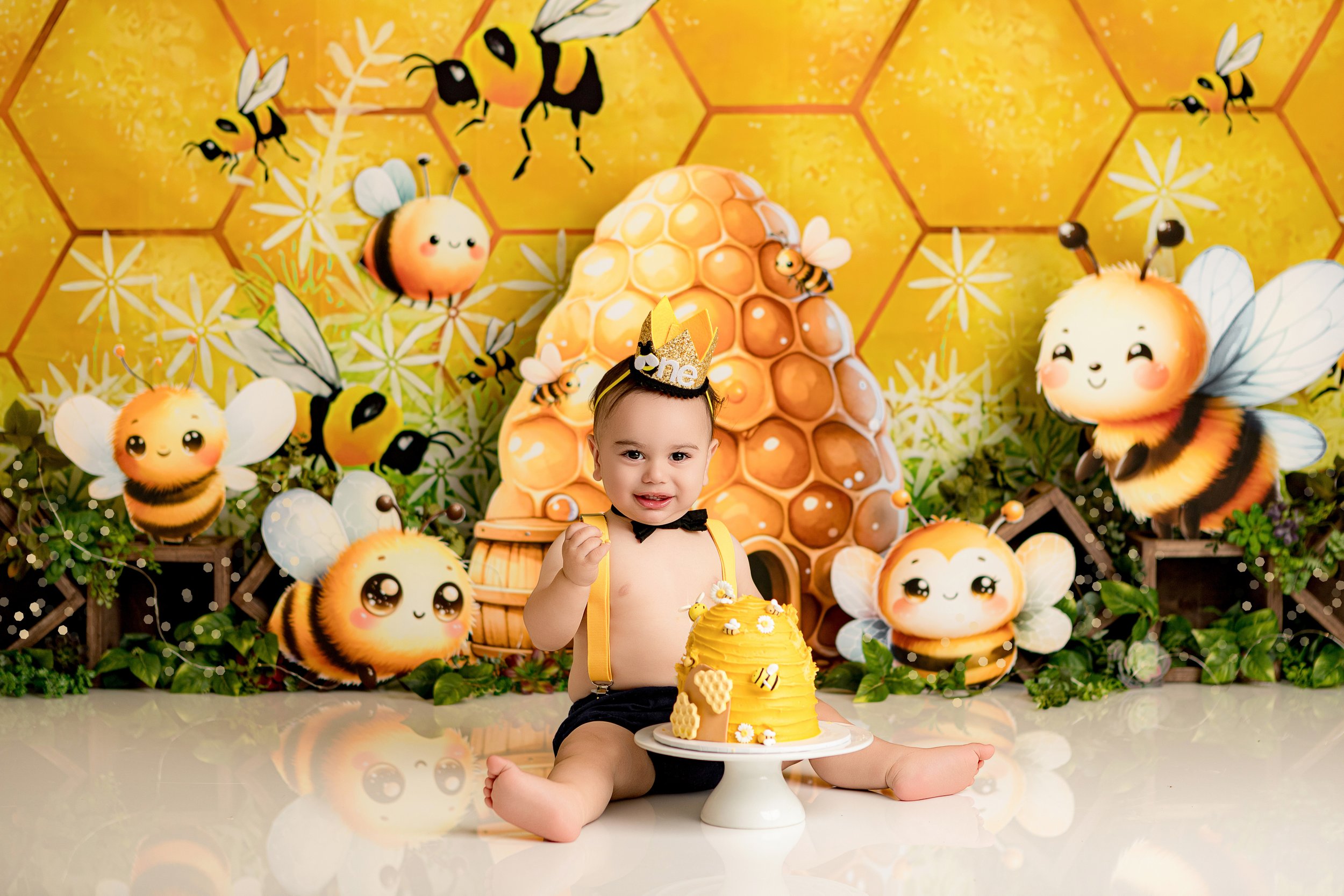 A toddler with a bee-themed party hat sitting on the floor in front of a honey bee-themed birthday cake, surrounded by honey bee and hive decorations. Family newborn photography Melbourne Cake Smash South Morang Photographer.