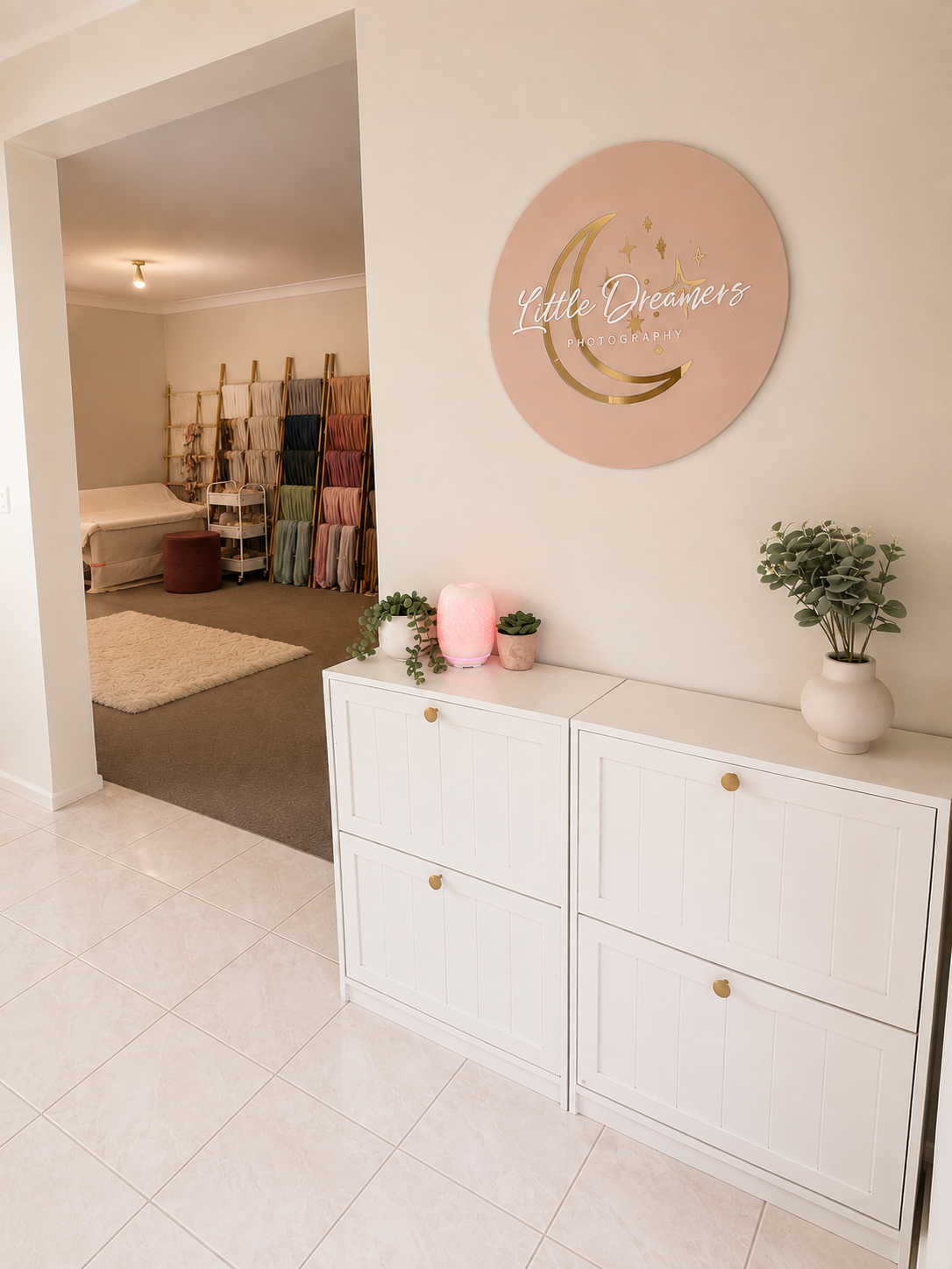 Interior of a photography studio with a sign that says 'Little Dreamers Photography,' a white cabinet with potted plants and a pink lamp, a fabric rack with colorful fabric swatches, and a cozy corner with a bed and a rug.