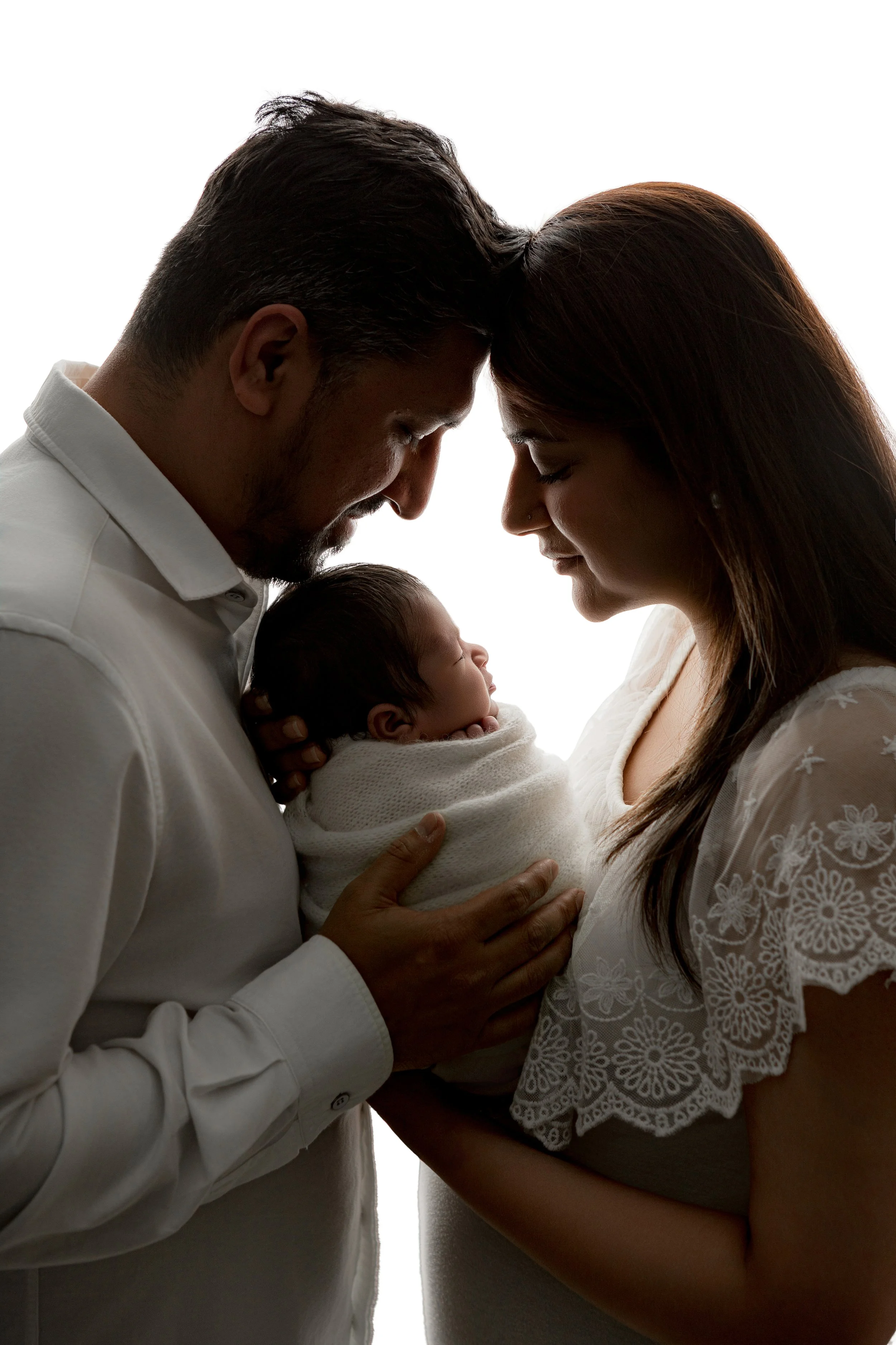 Silhouette image of a man and woman gently touching foreheads with a newborn baby.