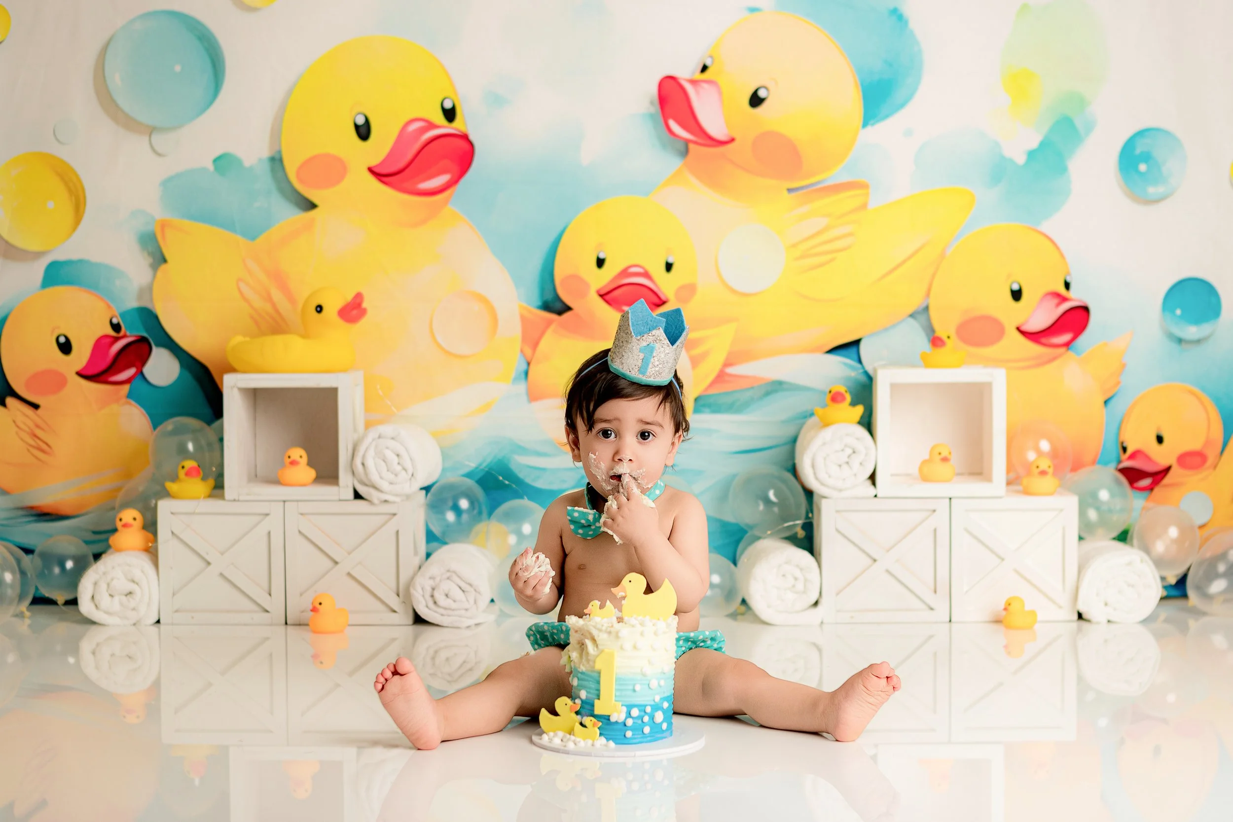 A young child celebrating a birthday with a rubber duck themed party, wearing a crown and a polka-dotted bow tie, sitting in front of a cake decorated with a number one candle, surrounded by rubber duck decorations and a colorful duck-themed backdrop.