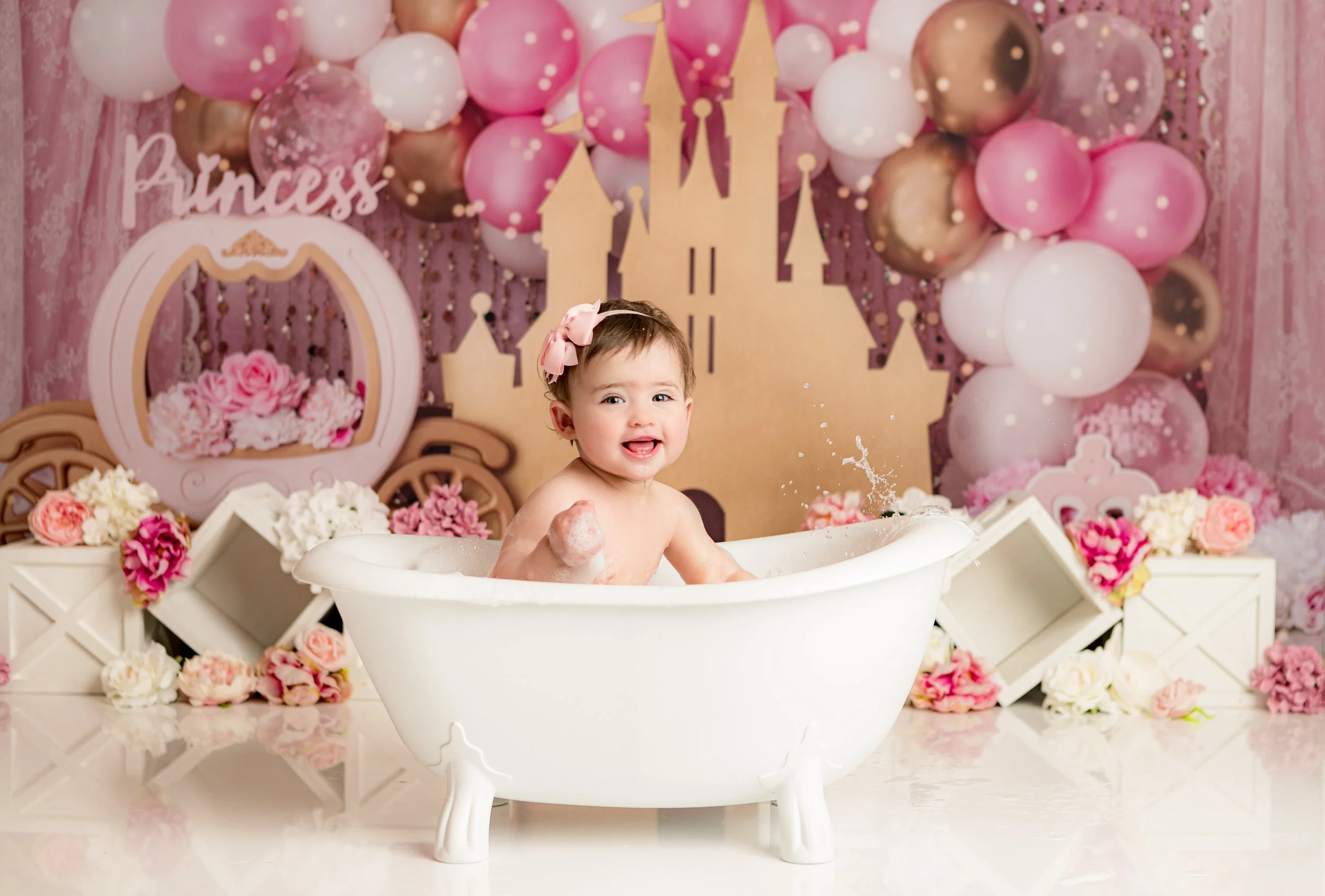 A young girl in a bathtub surrounded by pink, white, and brown balloons and pink flowers, with a castle backdrop and a "Princess" sign behind her. Cake Smash Photography Melbourne