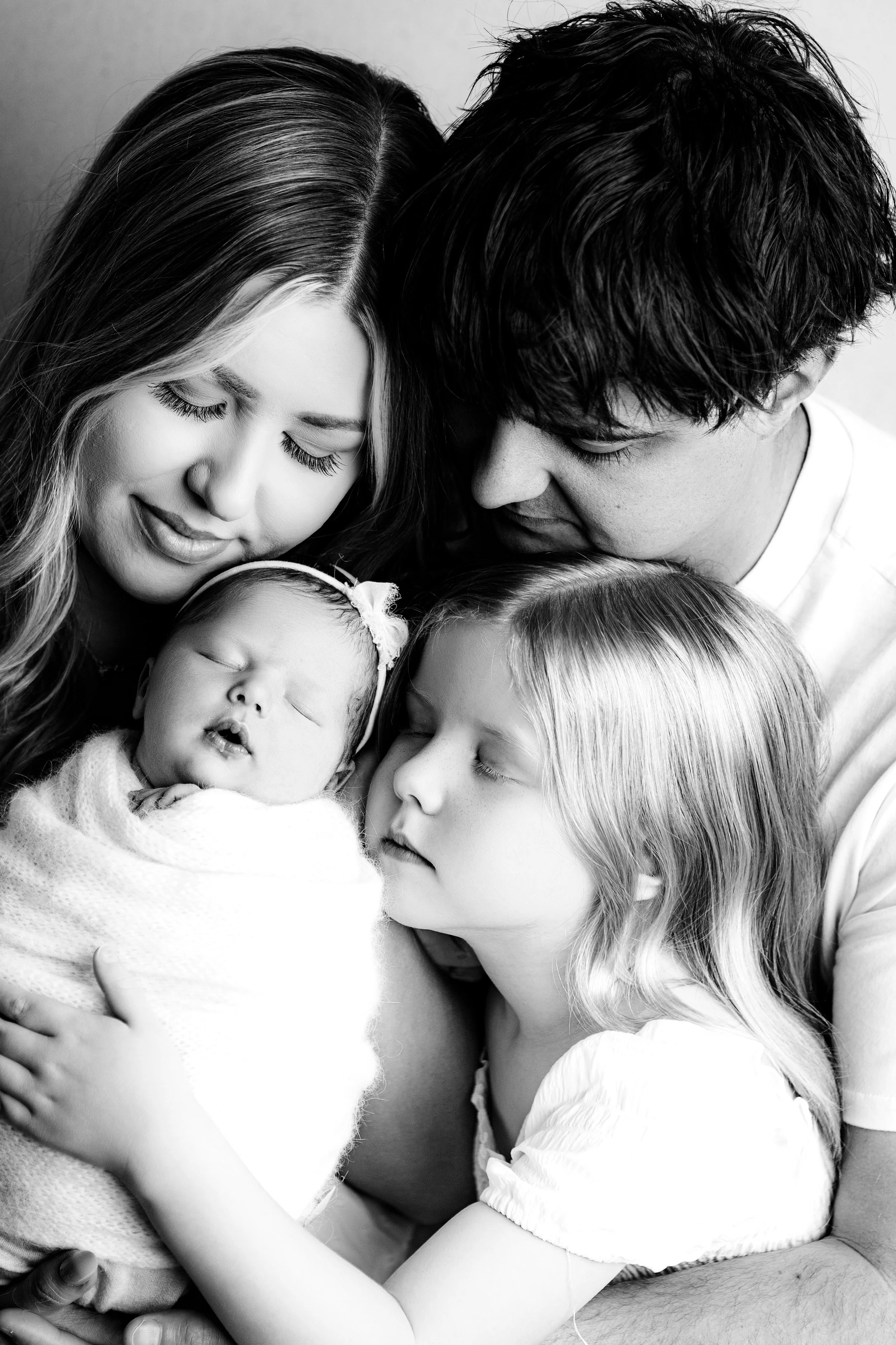 Black and white photo of a family, including a mother, father, and three young children, all cuddling together with closed eyes. Melbourne newborn photography South Morang