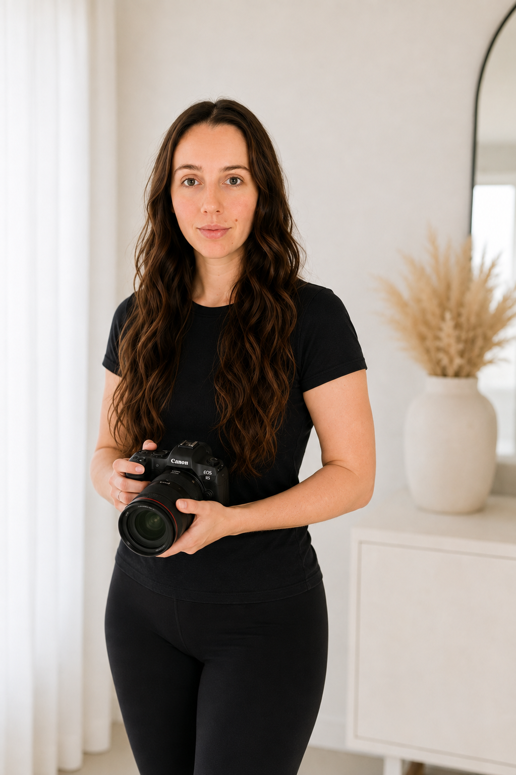 A woman with long wavy brown hair holding a Canon DSLR camera, standing indoors near a white cabinet with a potted plant in the background. Photographer Melbourne, South Morang.