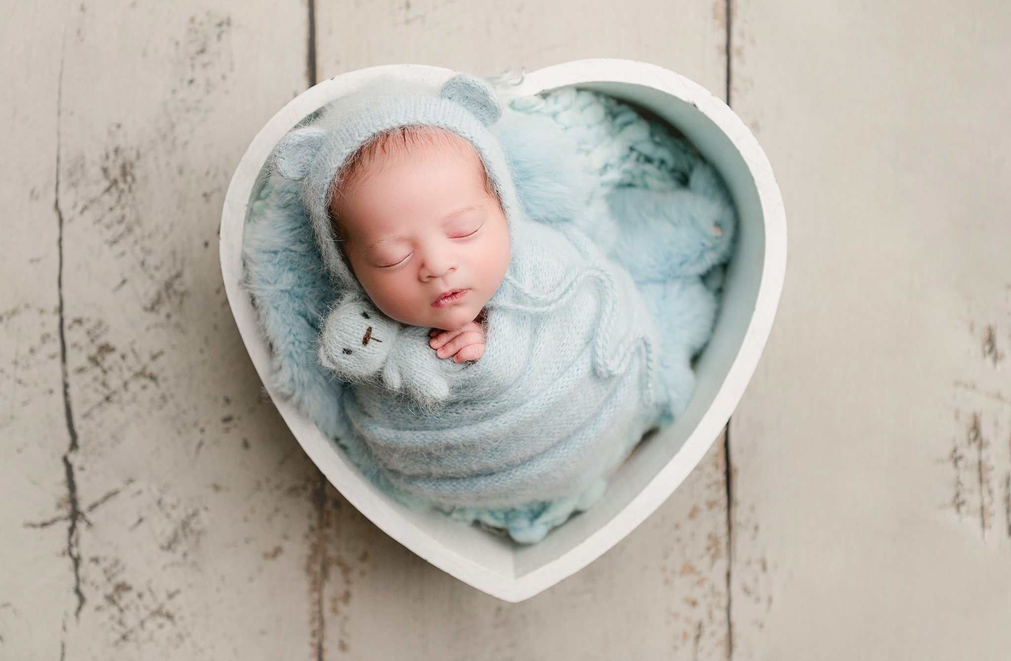 A sleeping baby wrapped in a fuzzy blue blanket and wearing a knit hat with bear ears, lying inside a heart-shaped box, on a rustic wooden floor. Melbourne newborn photography South Morang