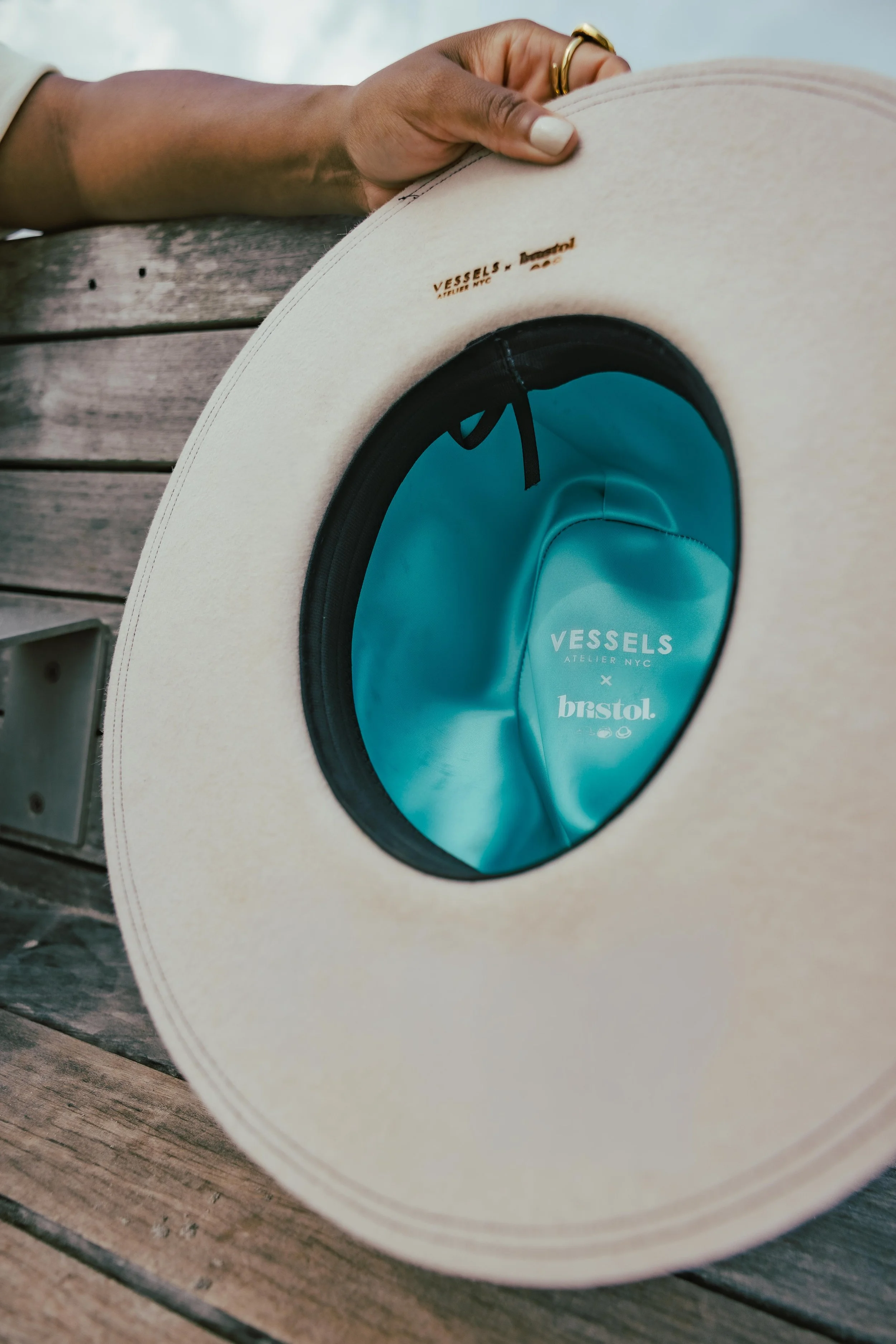 A beige wide-brimmed hat resting on a wooden surface, with the inside lining visible showing branding for VESSELS and bristol.