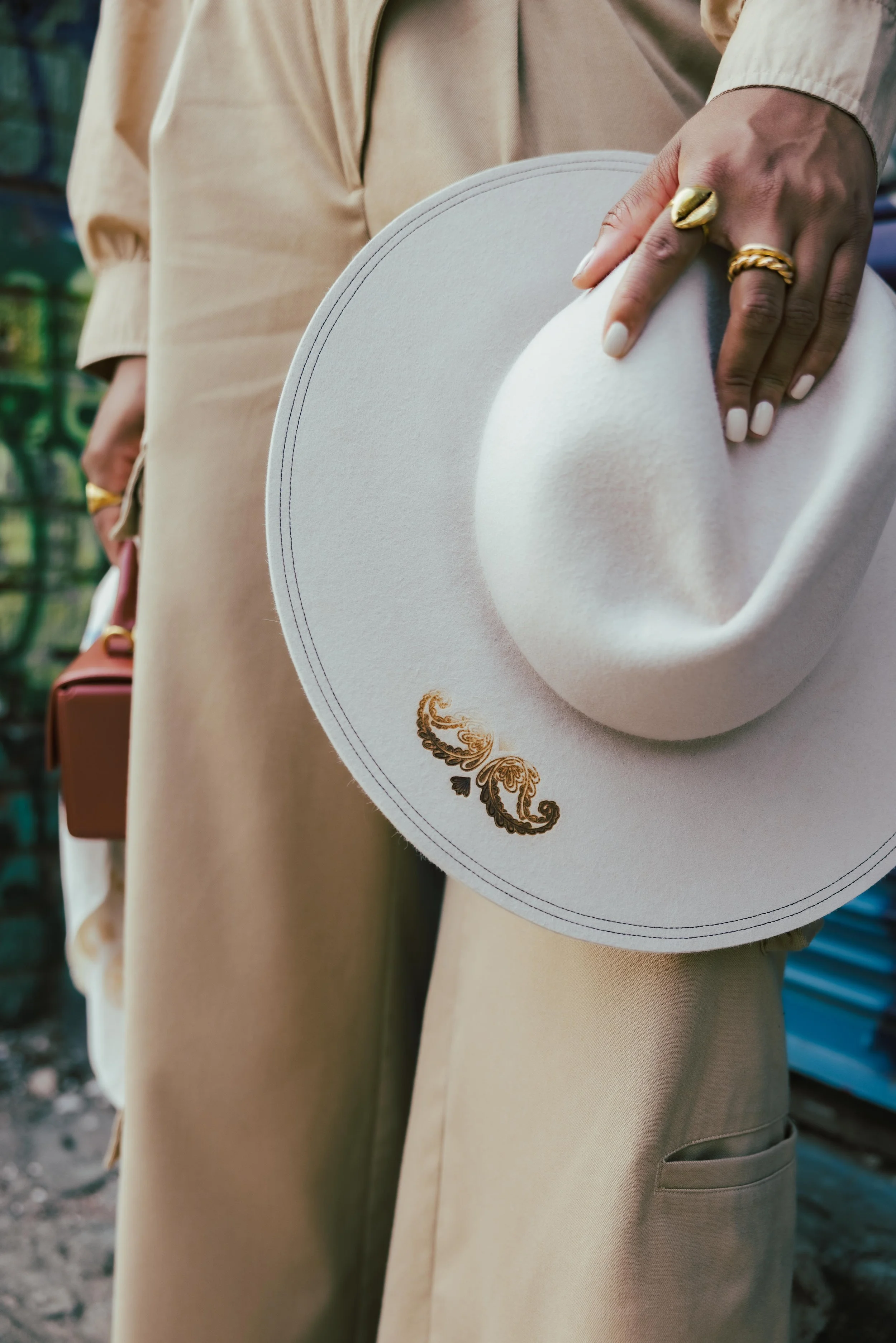 Person holding a white hat with gold embroidery, wearing beige clothing and gold rings, standing outdoors