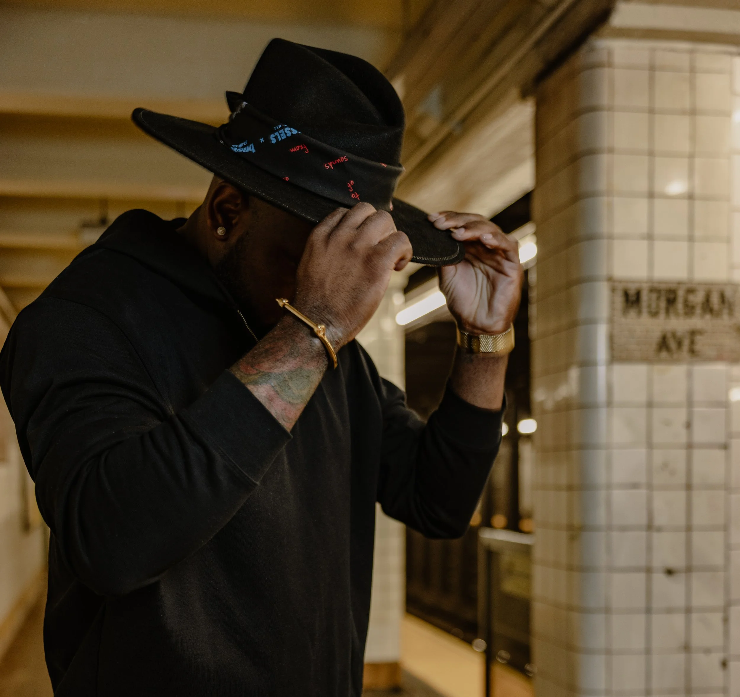 Man wearing a black hoodie and gold bracelet, adjusting his black fedora hat with red and blue accents in a subway station with white tiled walls.
