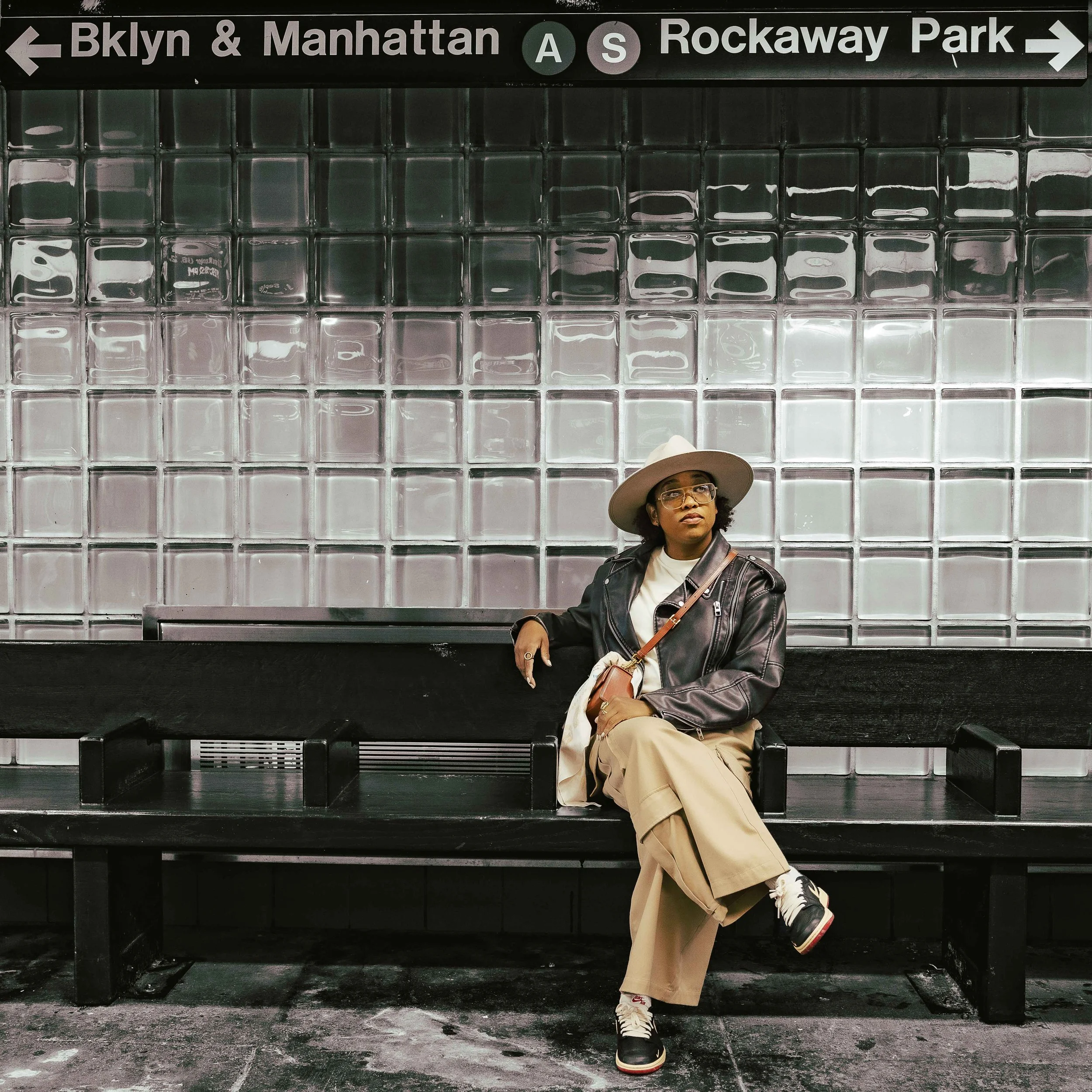 A woman sitting on a black bench in a subway station, wearing a wide-brimmed hat, glasses, a leather jacket, beige pants, and sneakers. Behind her is a wall of glass blocks and a sign indicating directions to Brooklyn & Manhattan and Rockaway Park, w