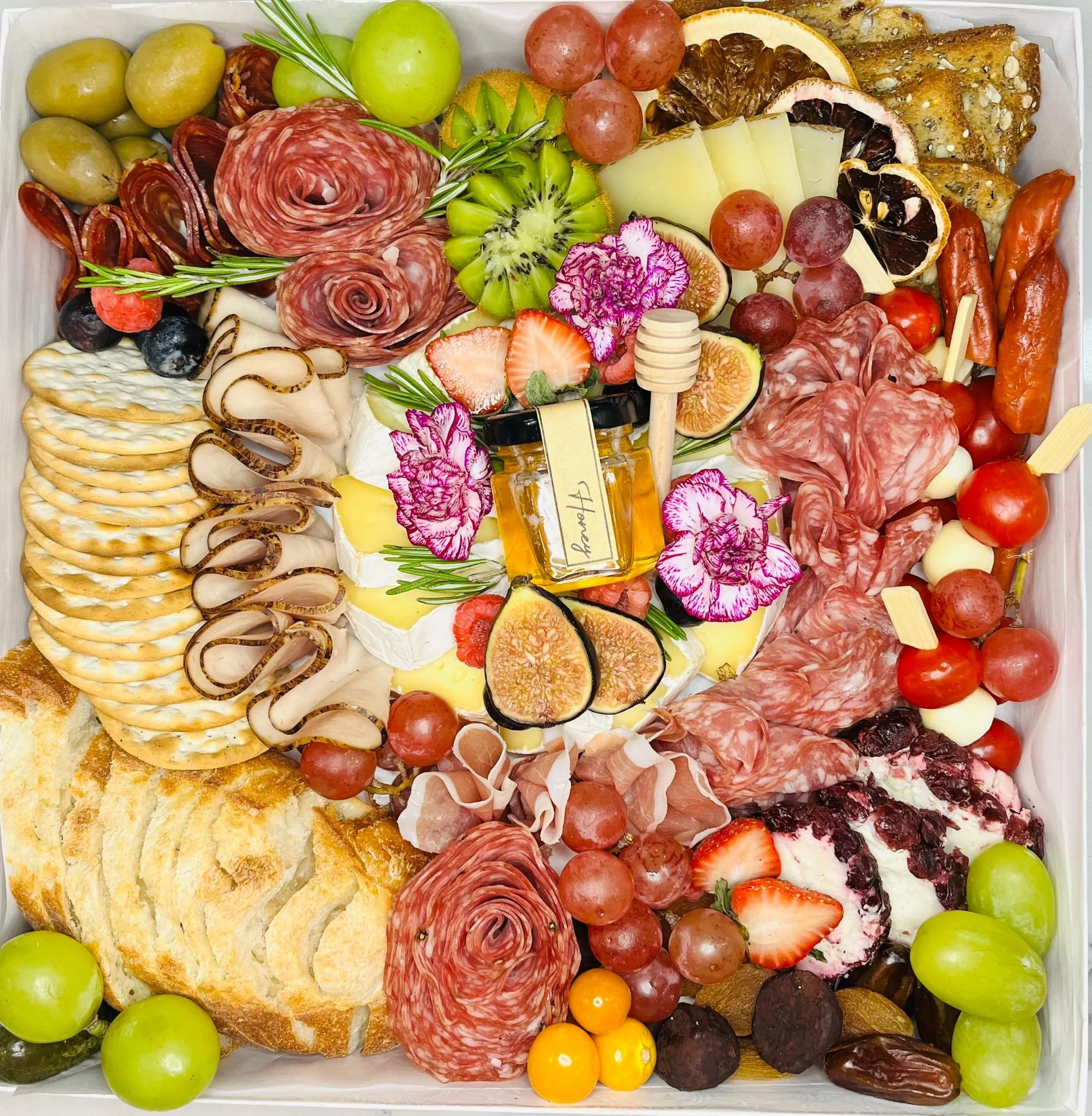 An assorted charcuterie board with salami, prosciutto, sliced bread, various cheeses, grapes, strawberries, figs, blueberries, and decorative edible flowers with honey at the center.