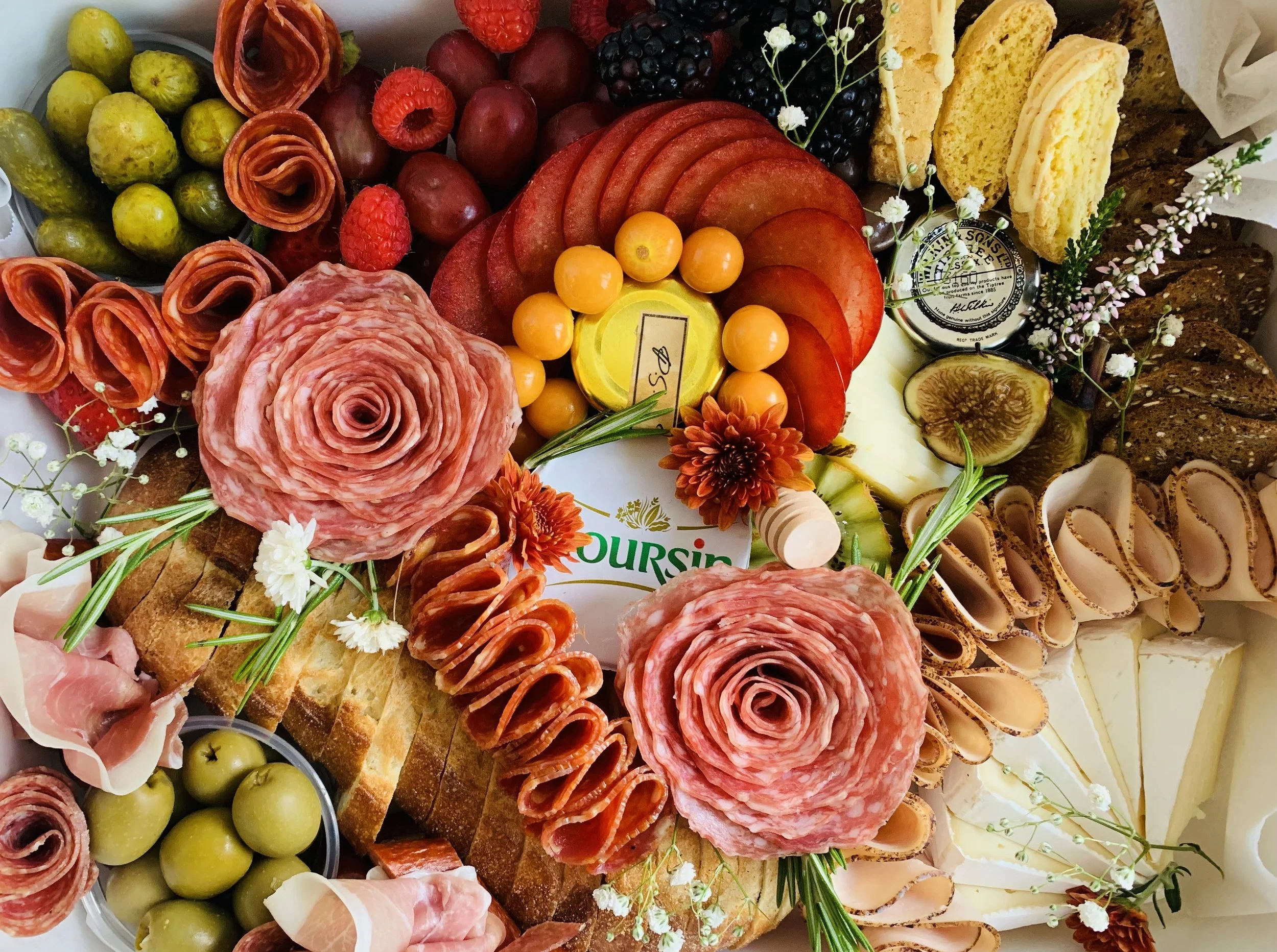 A variety of sliced meats, cheeses, grapes, berries, bread, and crackers arranged on a platter for a charcuterie board, decorated with small flowers and sprigs of herbs.