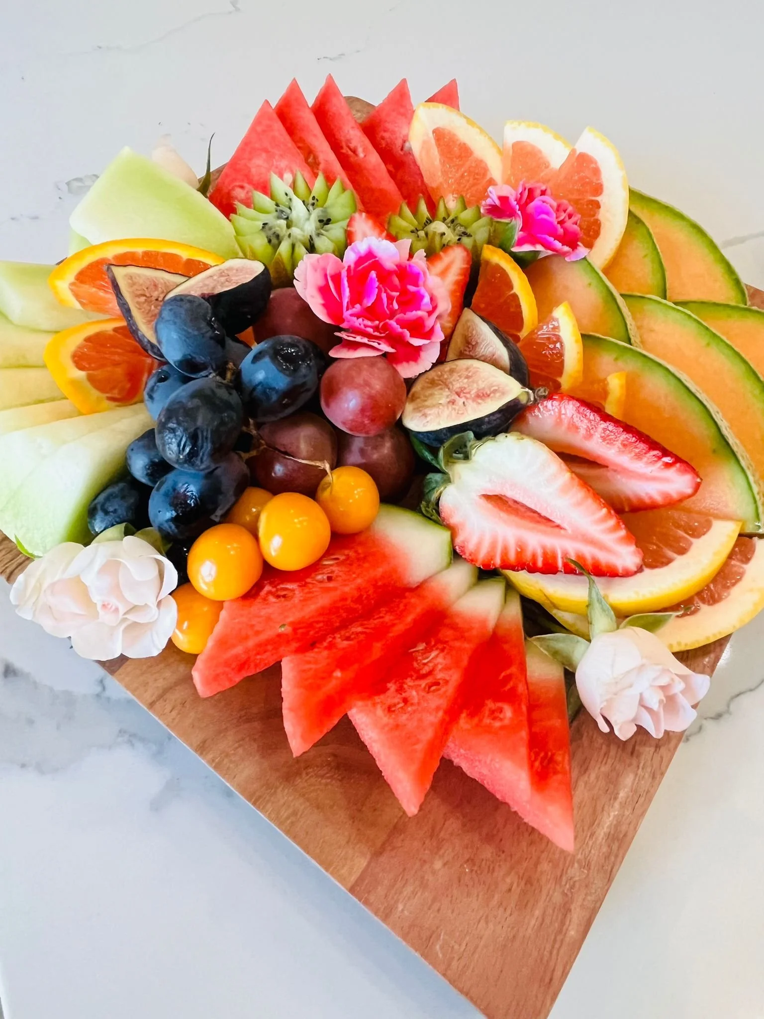 Assorted sliced and whole fruits, including watermelon, cantaloupe, honeydew, strawberries, blueberries, grapes, orange, grapefruits, and decorative flowers on a wooden tray.