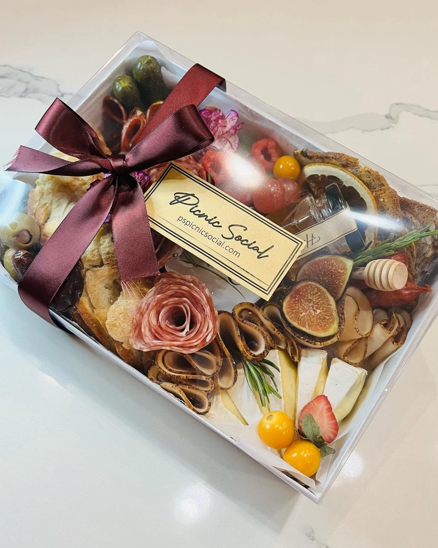Gift box labeled 'Picnic Social' containing assorted cheeses, figs, strawberries, grapes, cured meats, herbs, honey, and crackers, decorated with a purple ribbon.