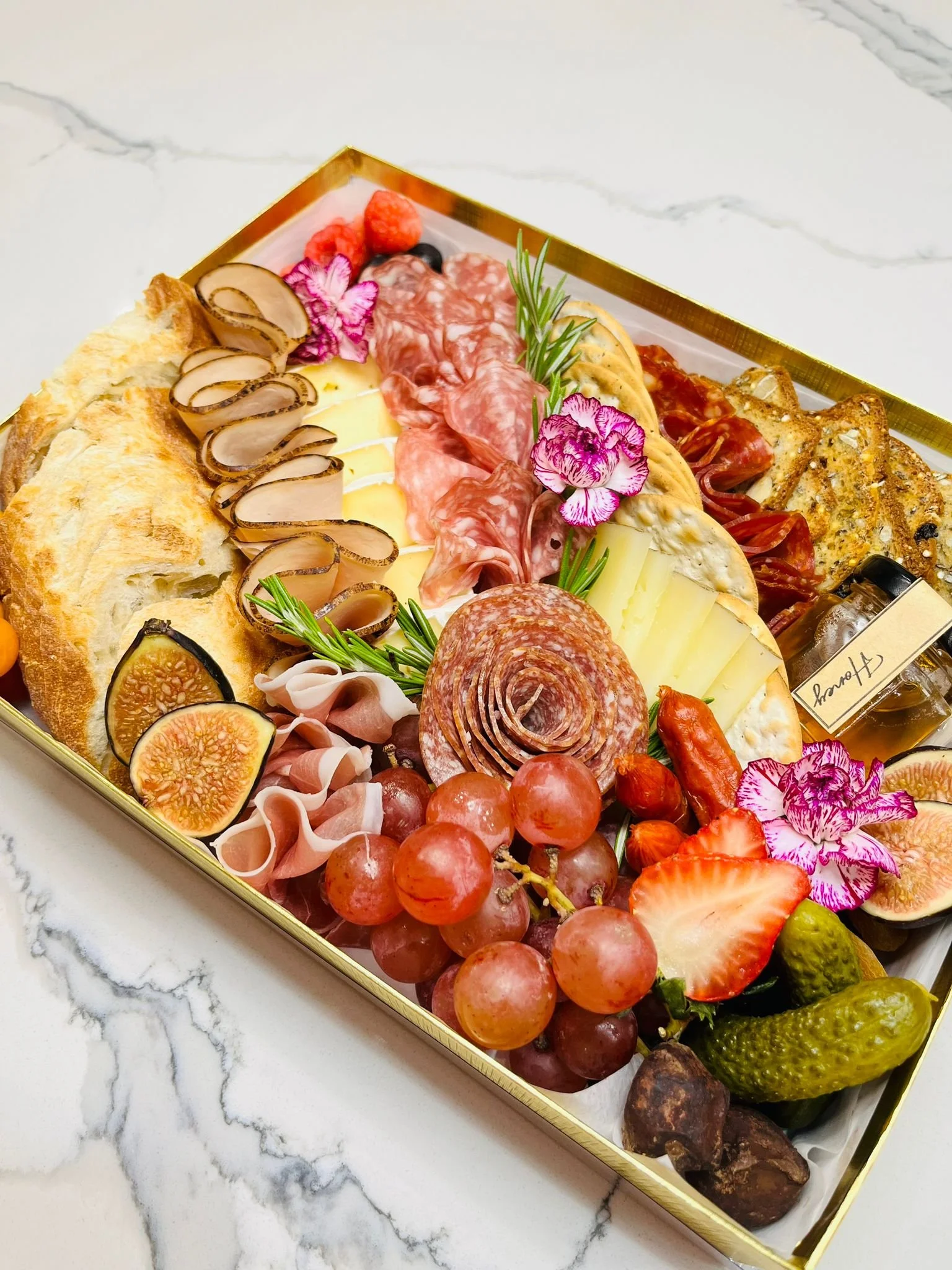 A gourmet charcuterie board with various cheeses, cured meats, fruits, bread, and garnishes arranged in a gold tray.