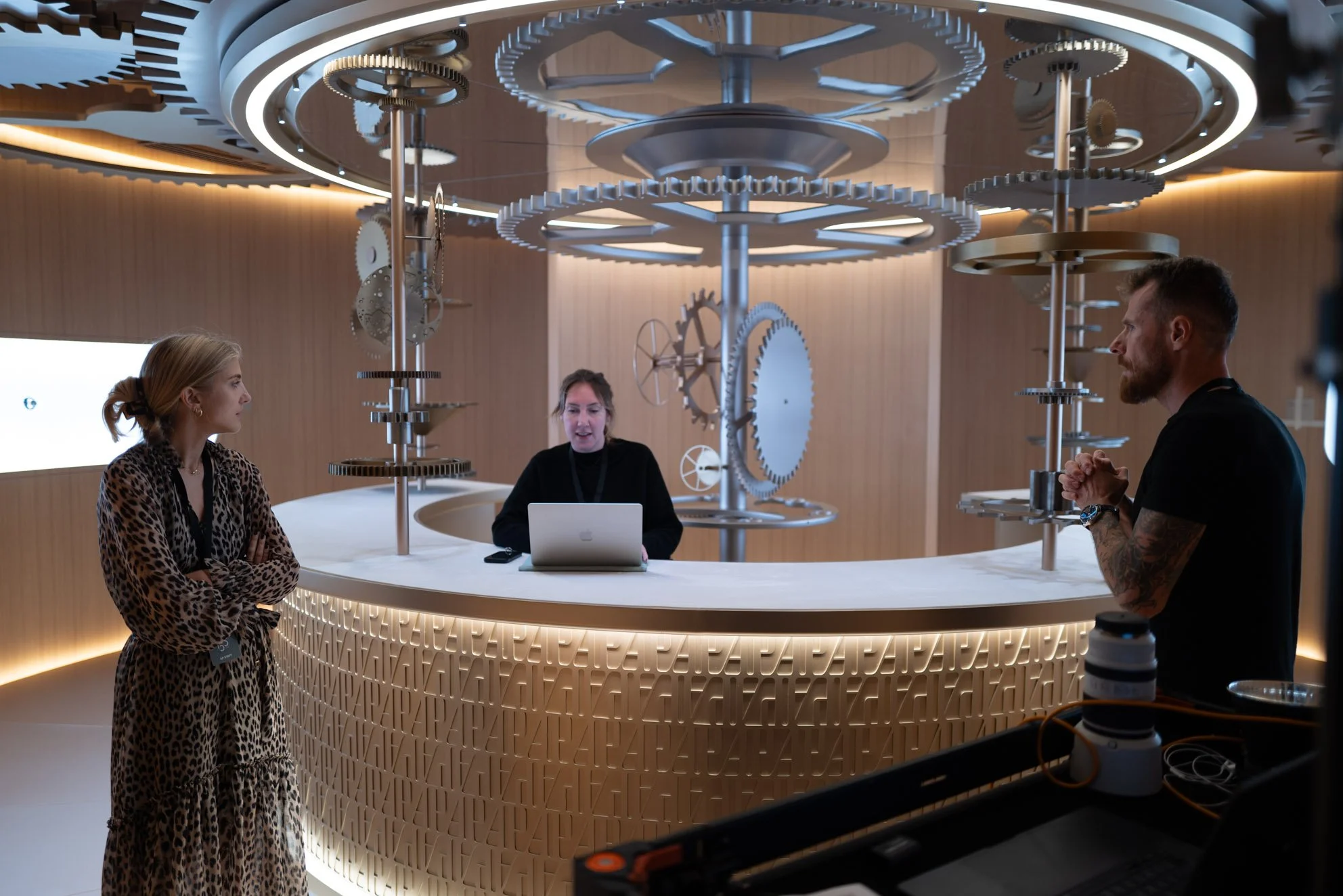 Three people in a modern room with a large circular desk and mechanical gears and cogs in the ceiling. One woman is standing with arms crossed, another woman is sitting behind the desk with a laptop, and a man with tattoos is standing on the other si