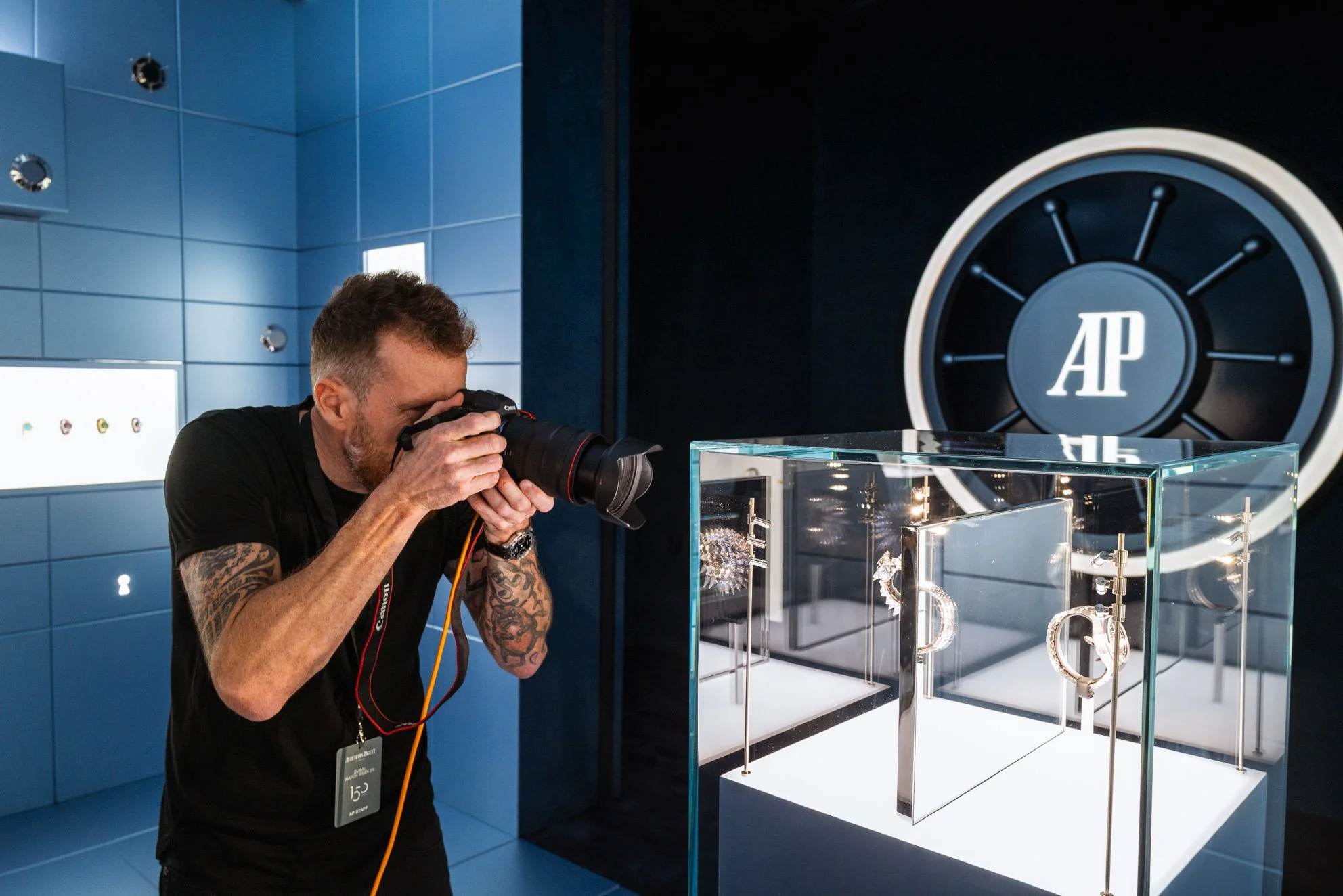 A man taking a photograph with a professional camera in a jewelry display room. The display contains jewelry pieces like earrings and necklaces inside a glass case. The background features a large vault door with the initials"AP" on it, and blue wall
