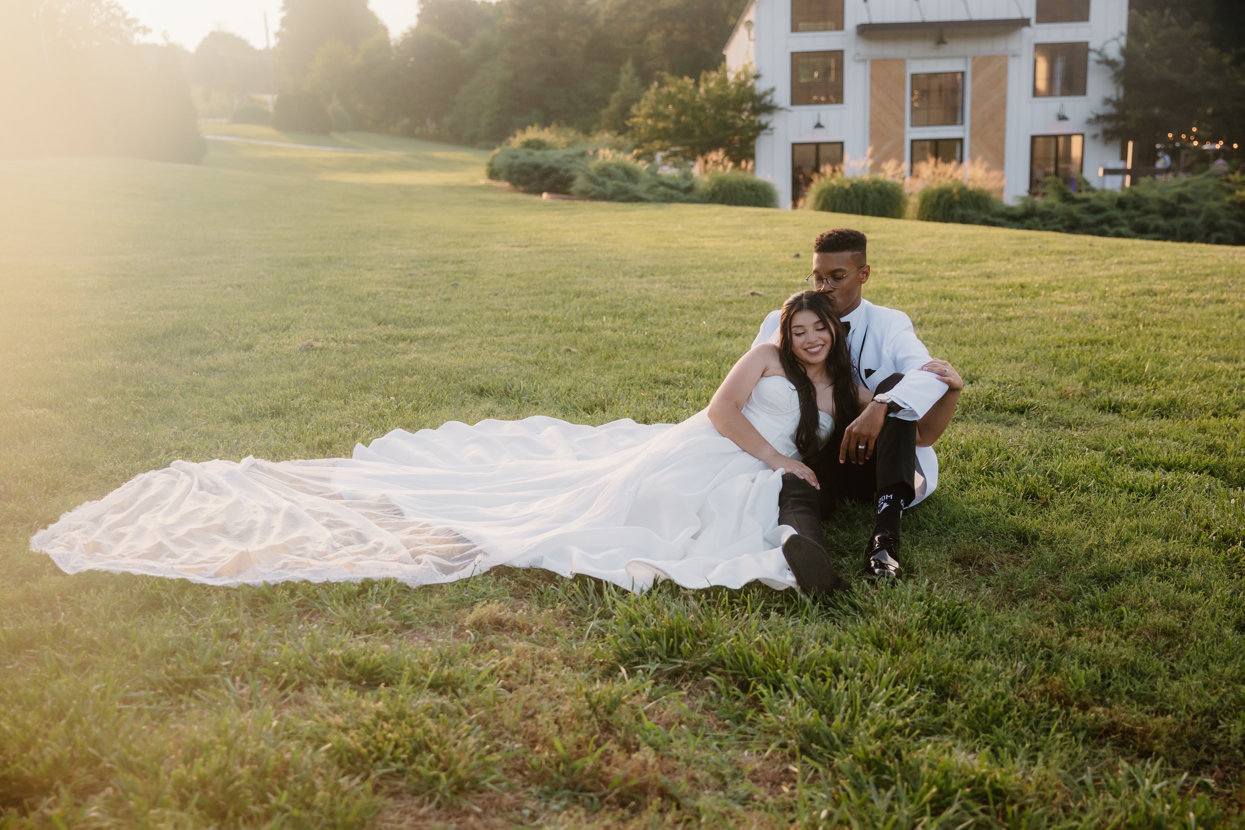 A couple sitting together in a field at golden hour, the bride's train spread out across the grass