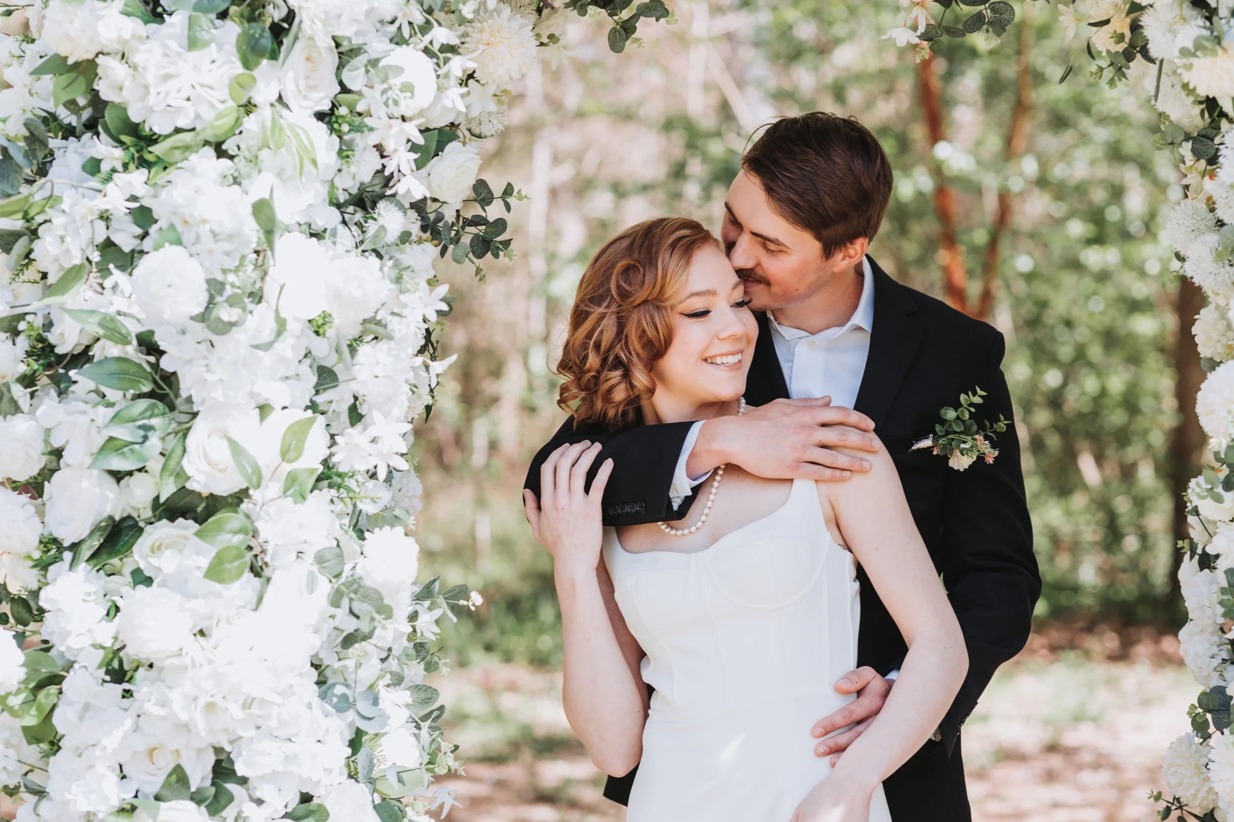 Bride and groom embracing beside a wall of white flowers at The Grand Ivy Point in Suwanee, GA