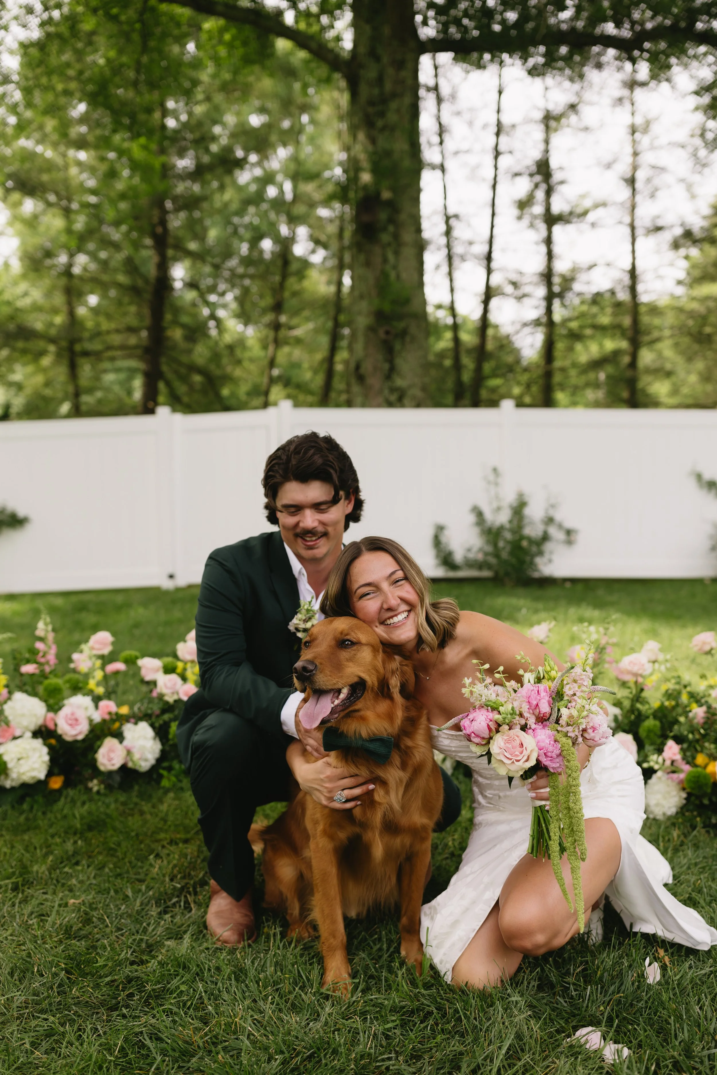 A happy couple in wedding attire with their dog on a grassy lawn, surrounded by pink and white flowers, in a backyard with trees and a white fence.