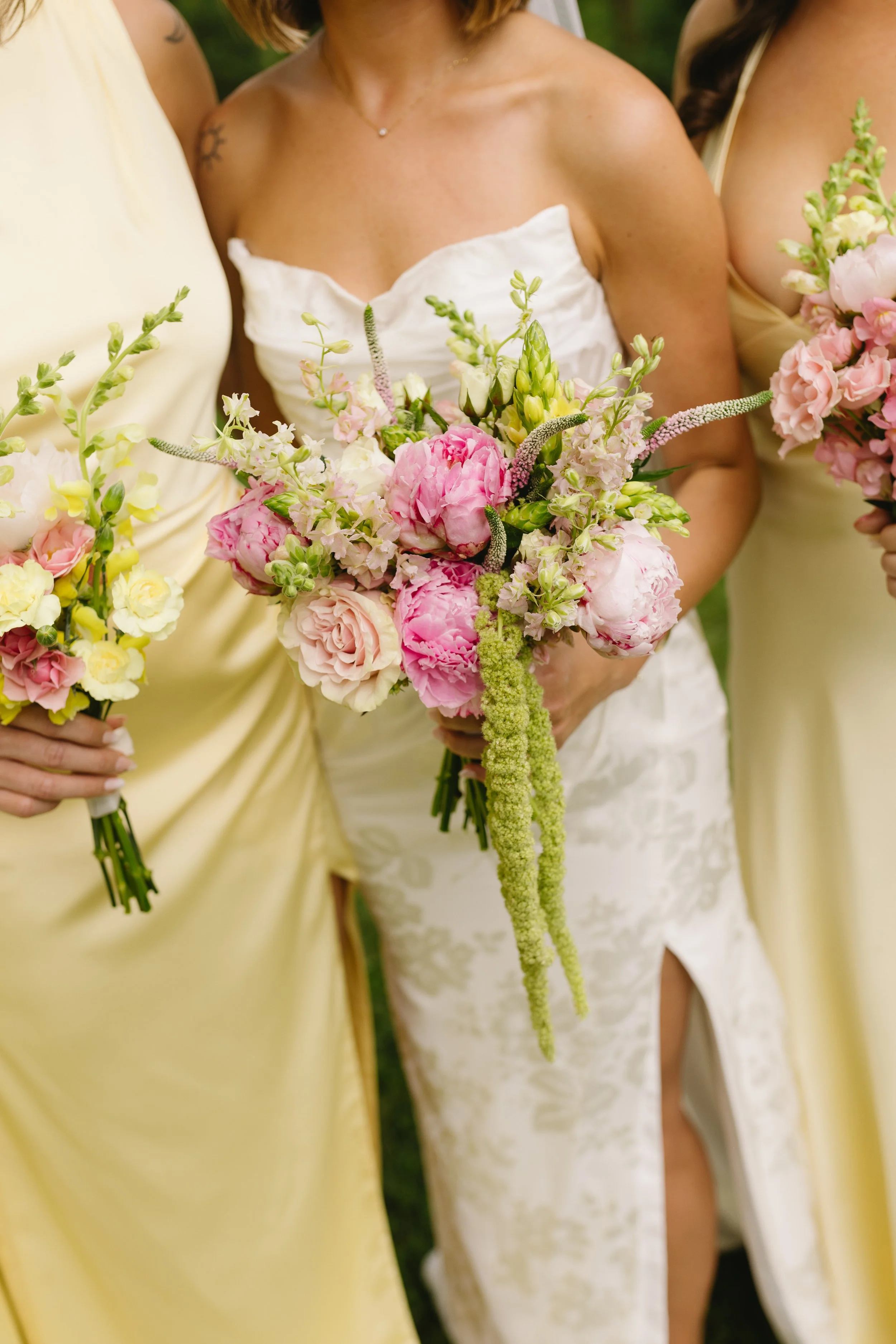 Close-up of three women in wedding and bridesmaid dresses holding pink and cream floral bouquets during a wedding ceremony.