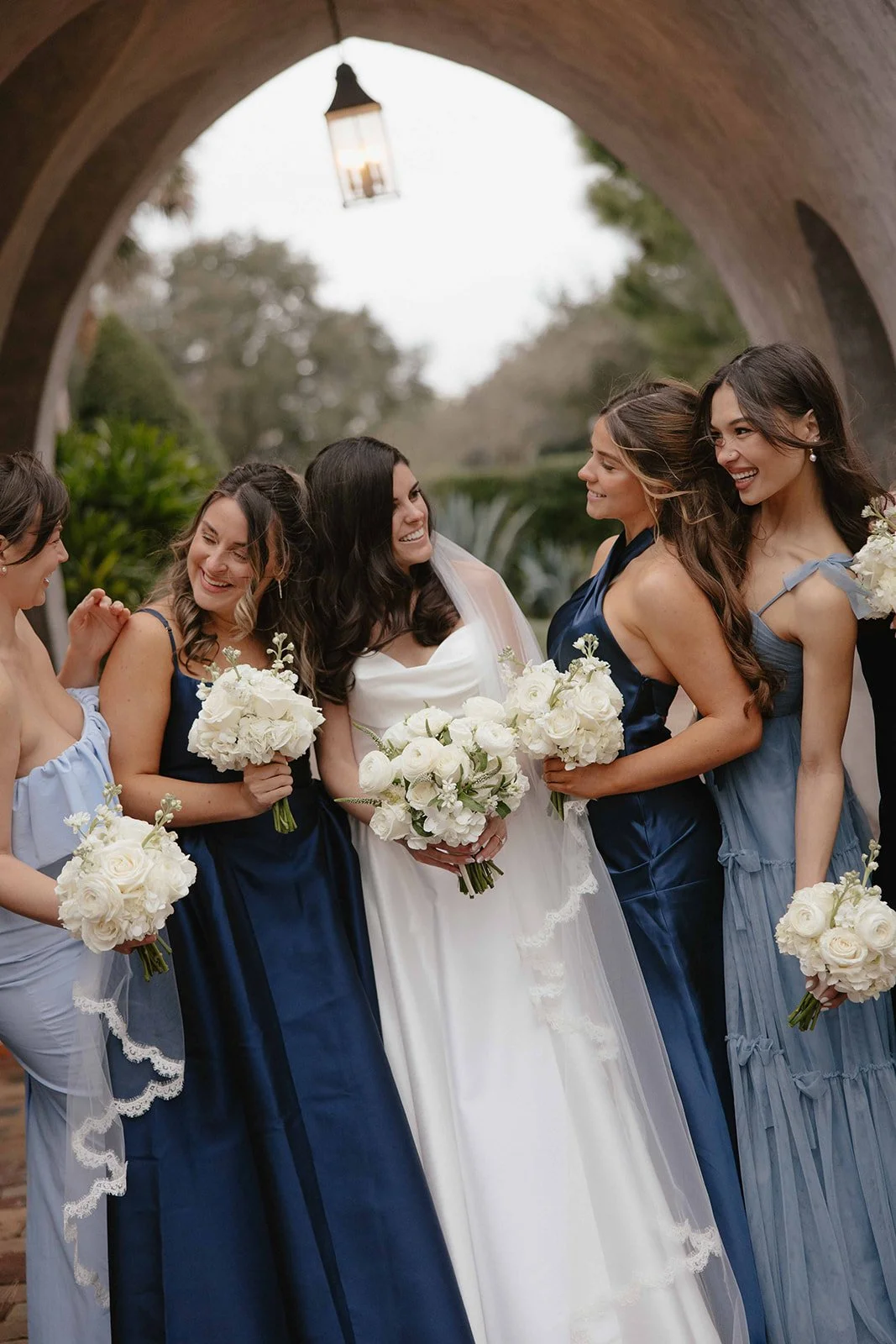A bride and four bridesmaids standing together outdoors beneath an archway, all holding bouquets of white flowers and smiling.