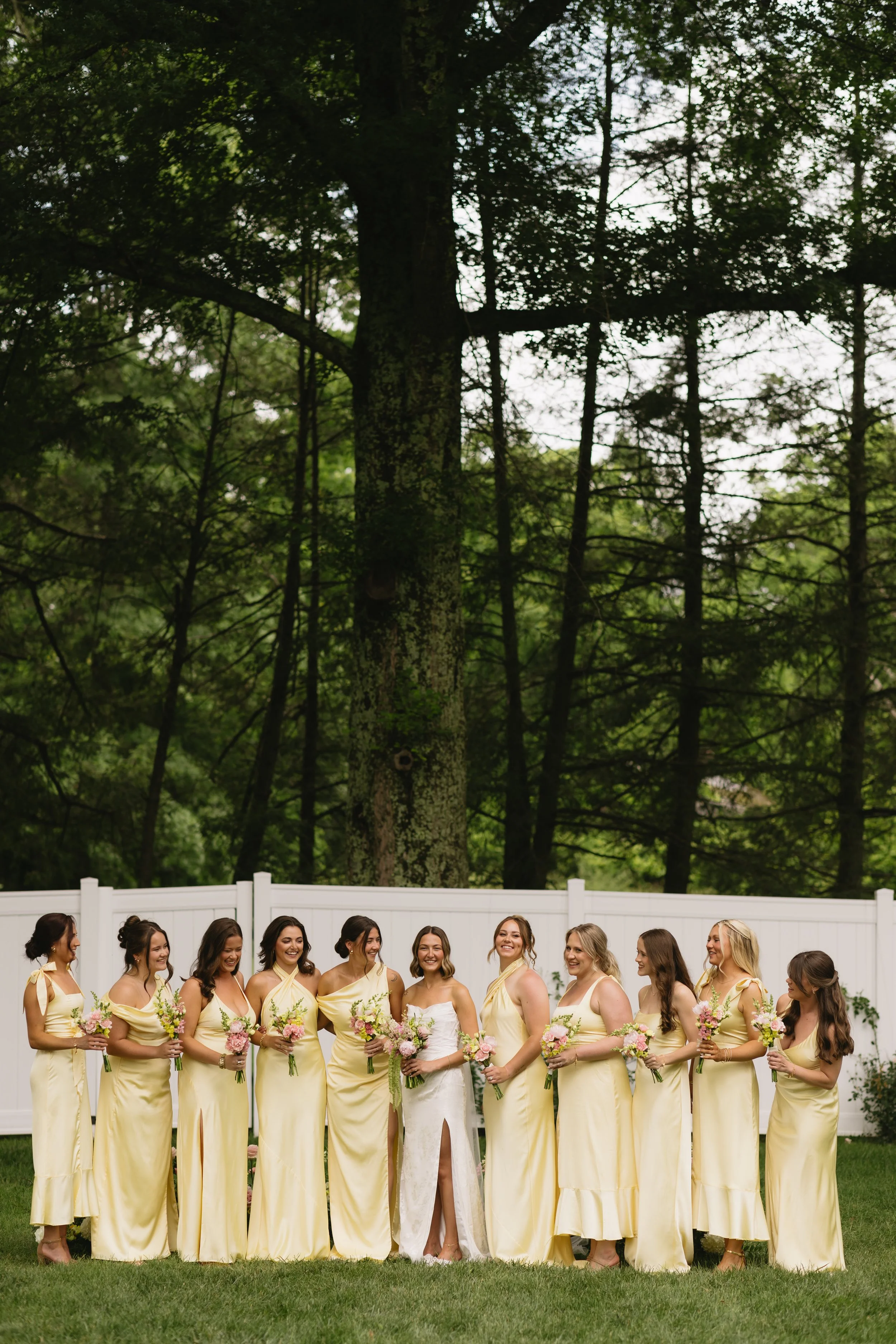 Bride in a white dress with a slit, standing among her bridesmaids who are all wearing yellow dresses, outdoors in front of a white fence and tall trees, holding pink and white bouquets.