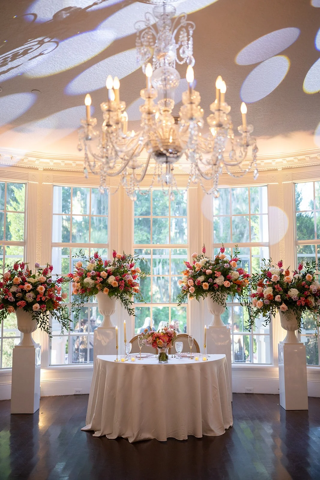 Elegant wedding reception setup with a round table draped in a white tablecloth, floral centerpieces in pink and white, surrounded by large floral arrangements in white vases, large windows with trees outside, and a crystal chandelier casting pattern