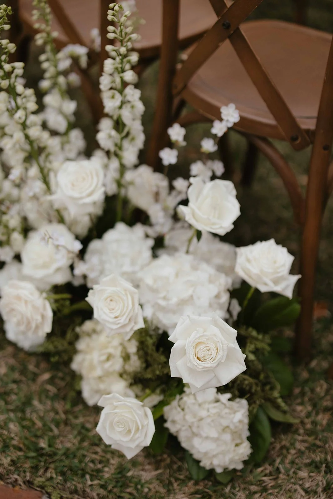 A floral arrangement with white roses, hydrangeas, and small white flowers next to a wooden chair at an outdoor event.