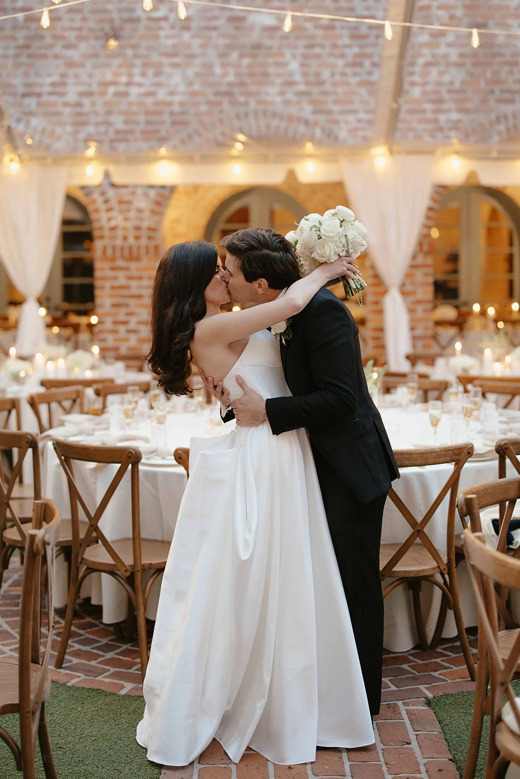 A bride and groom kiss at their wedding reception, with the bride holding a bouquet of white roses. The event is decorated with string lights and white drapery in a brick-walled venue.
