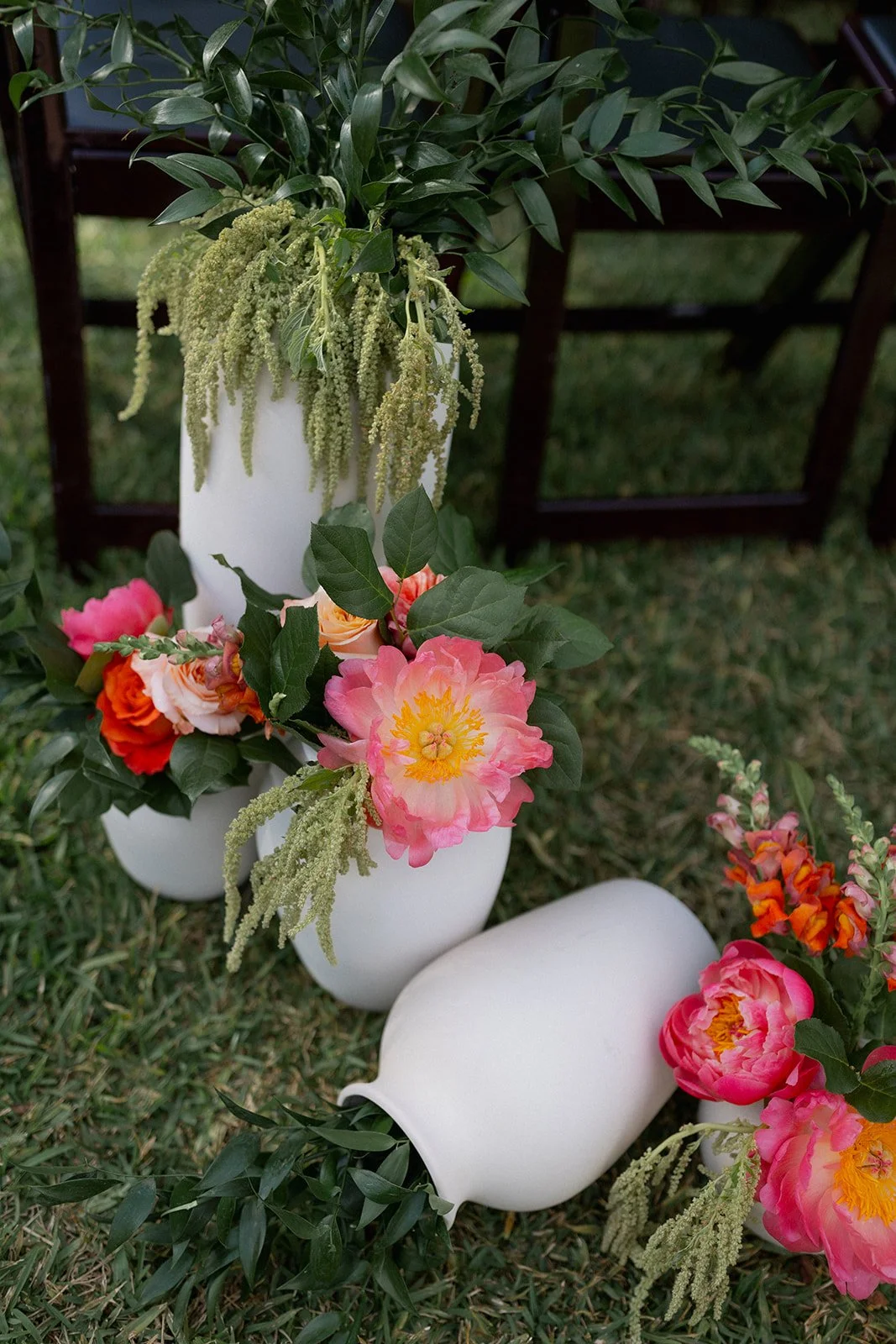 Flower arrangements with pink, orange, and peach peonies, greenery, and white vases on grass, with wooden chairs in the background.