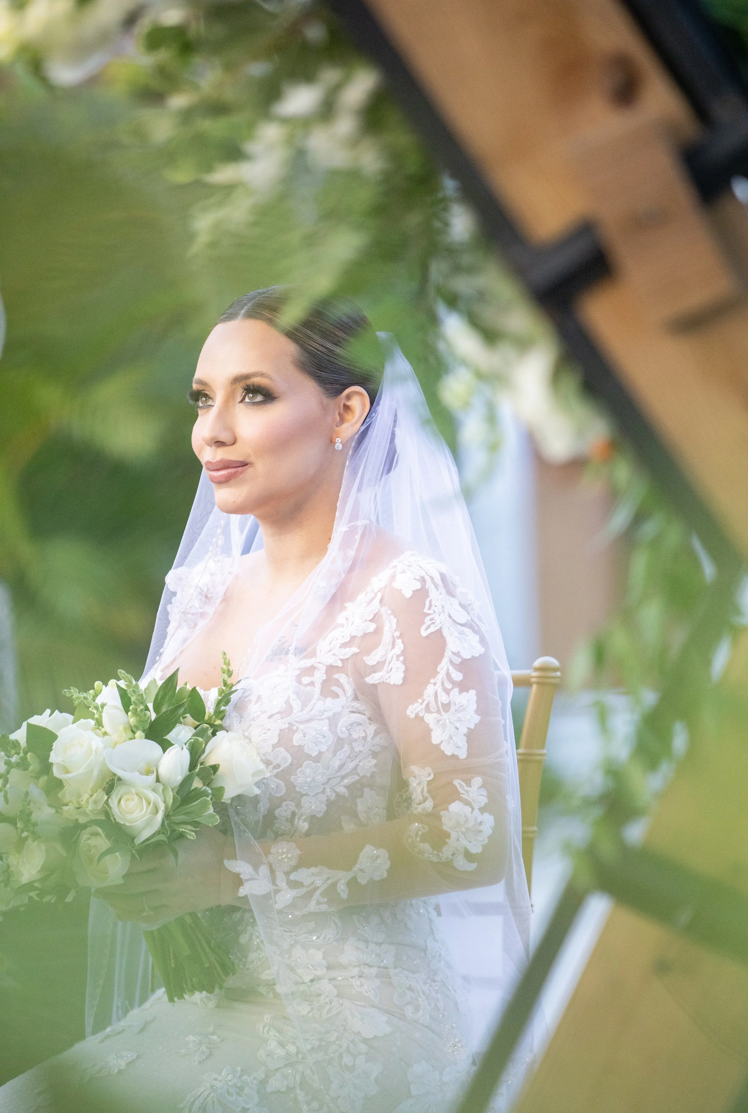 A bride dressed in a white lace wedding gown and veil, holding a bouquet of white roses and greenery, sitting on a gold chair with a wooden and green leafy background.