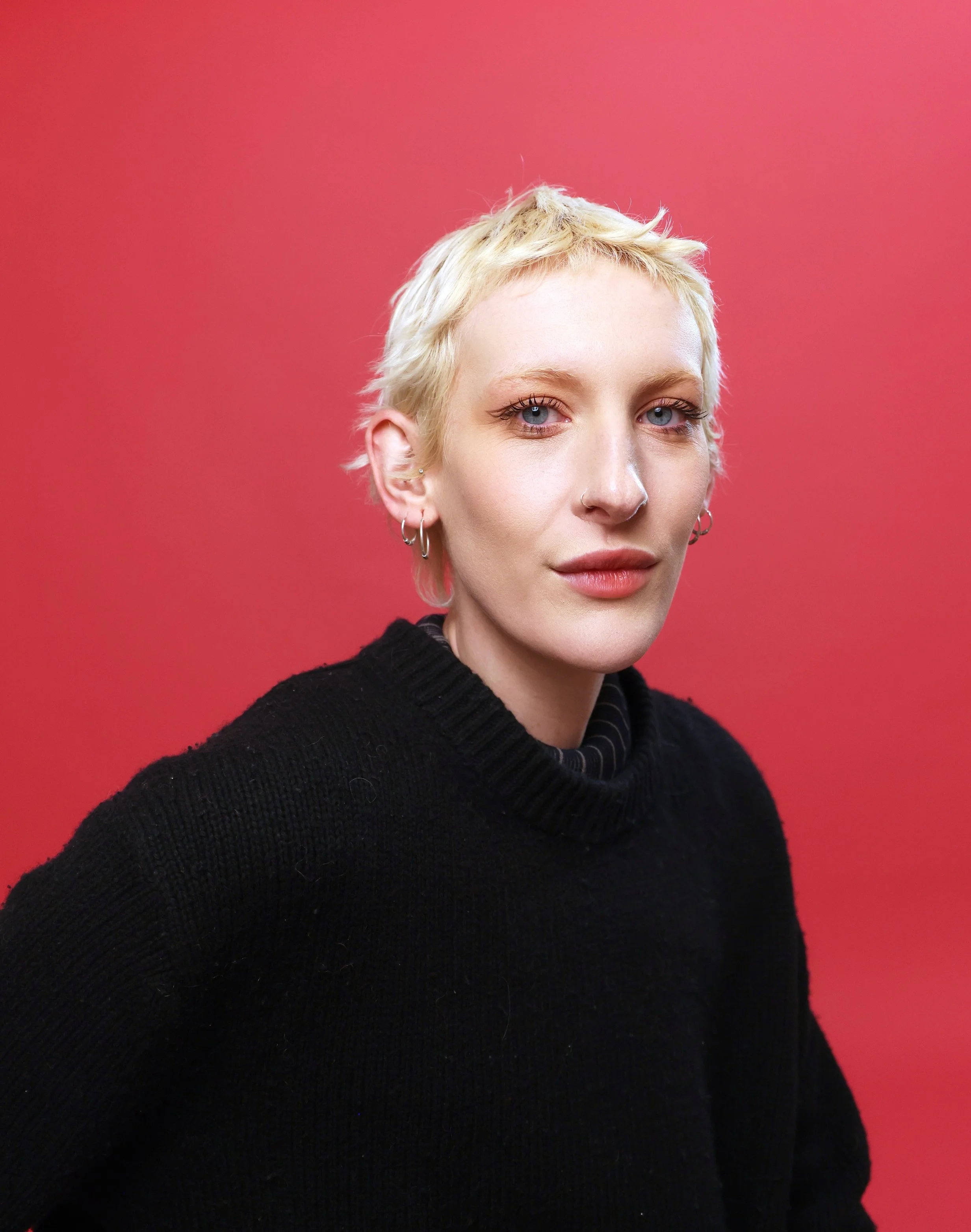 Portrait of a young woman with short blonde hair and multiple earrings, wearing a black sweater, against a pink background.