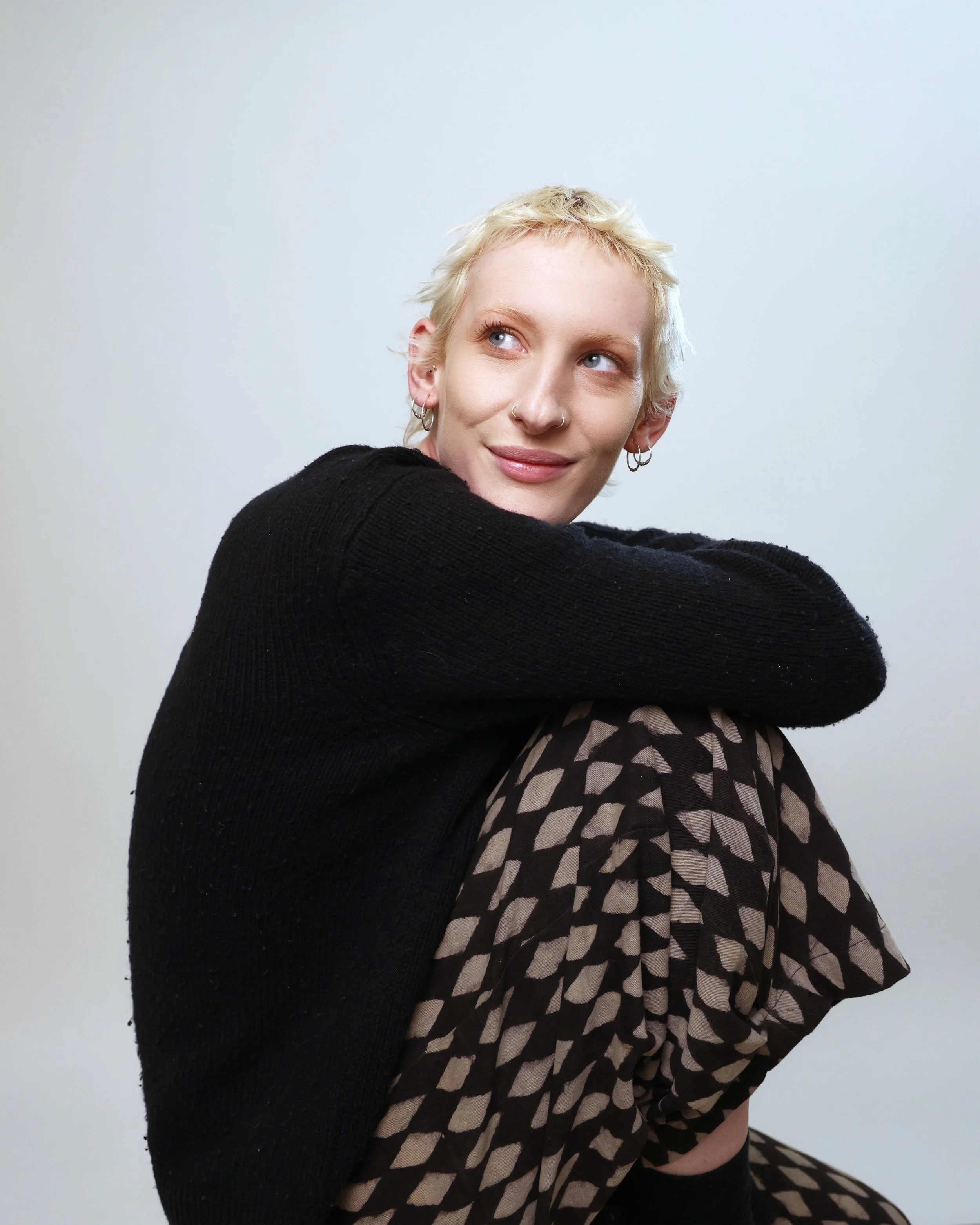 Young woman with short blonde hair and multiple earrings, sitting against a plain light background, wearing a black sweater and patterned pants, looking to her left with a slight smile.