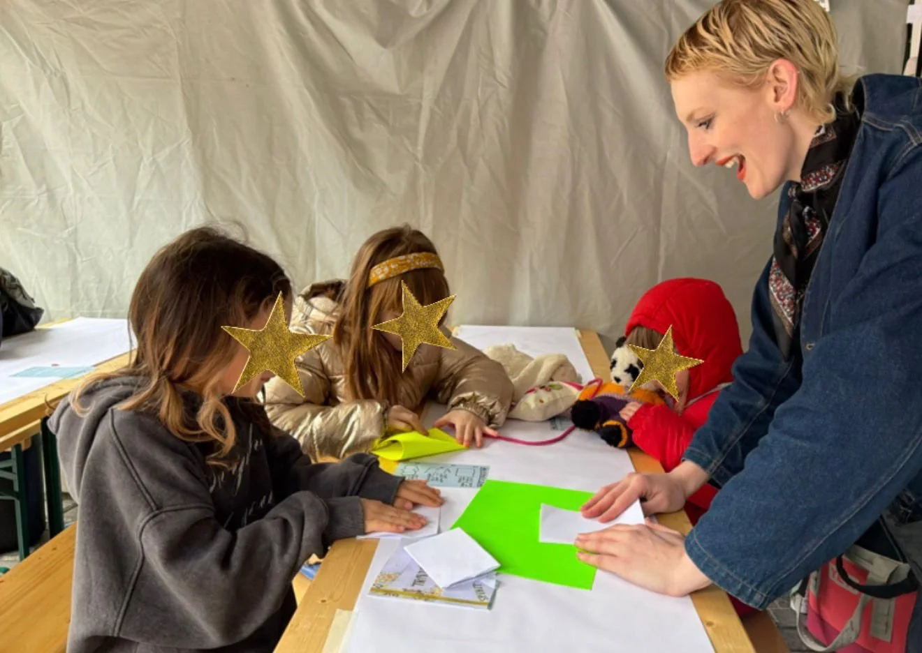 blonde hair, wearing a denim jacket, leaning over a table, engaging with three children whose faces are obscured by gold star stickers, inside a tent or temporary shelter with a beige backdrop. The children are dressed warmly, one in a red hoodie and another in a gold jacket, with papers and colorful materials on the table.