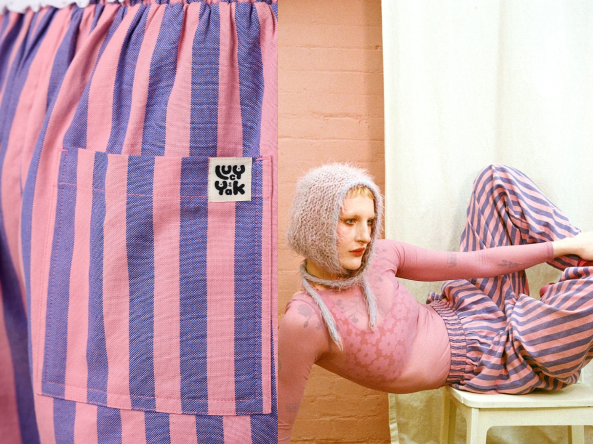 Close-up of pink and purple striped fabric with a small pocket and a label. A young woman with curly hair, wearing a fuzzy hooded head covering, pink long sleeve top, and striped pants, is reclining against a wall with her legs resting on a chair, in a room with peach-colored and white walls.