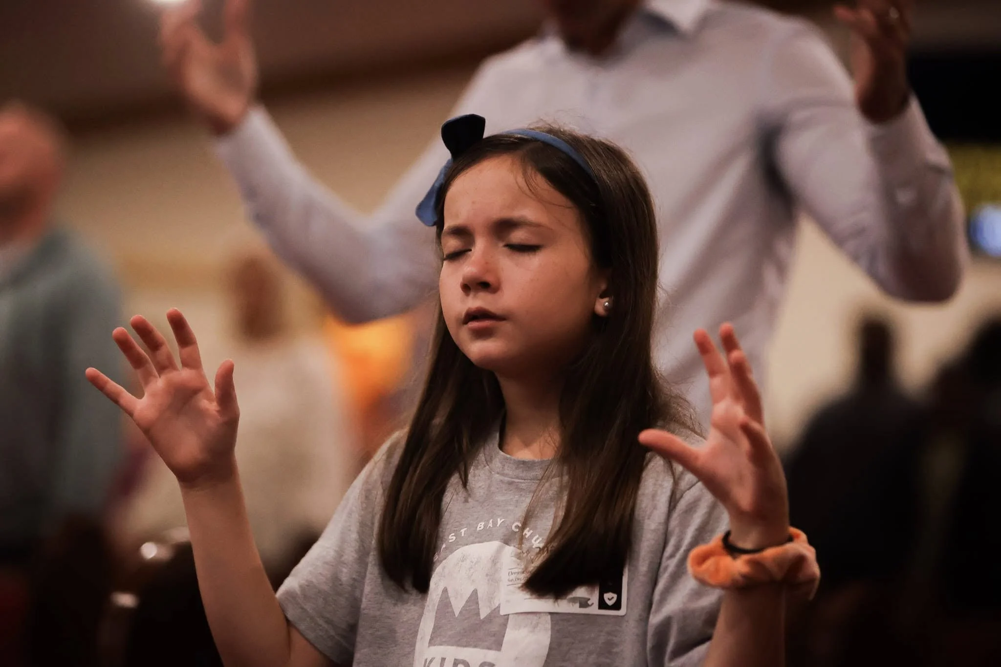 A young girl holding a sign that reads 'Follow Me To...' with a logo for 'Kids' in a church or conference hall, with people seated and a speaker on stage in the background.