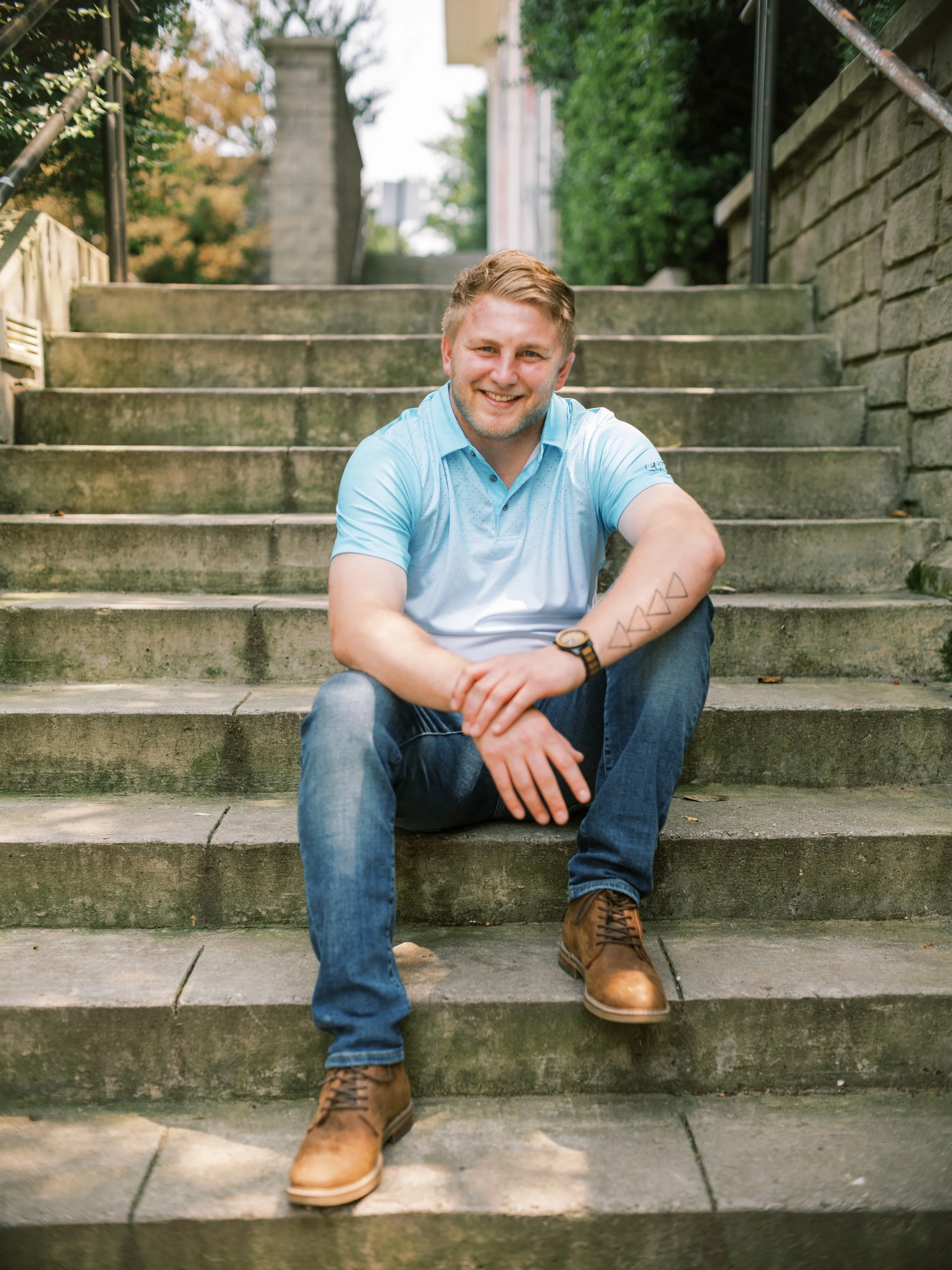A smiling young man with light brown hair, a light beard, wearing a light blue polo shirt, jeans, and brown boots, sitting on outdoor concrete steps surrounded by greenery.