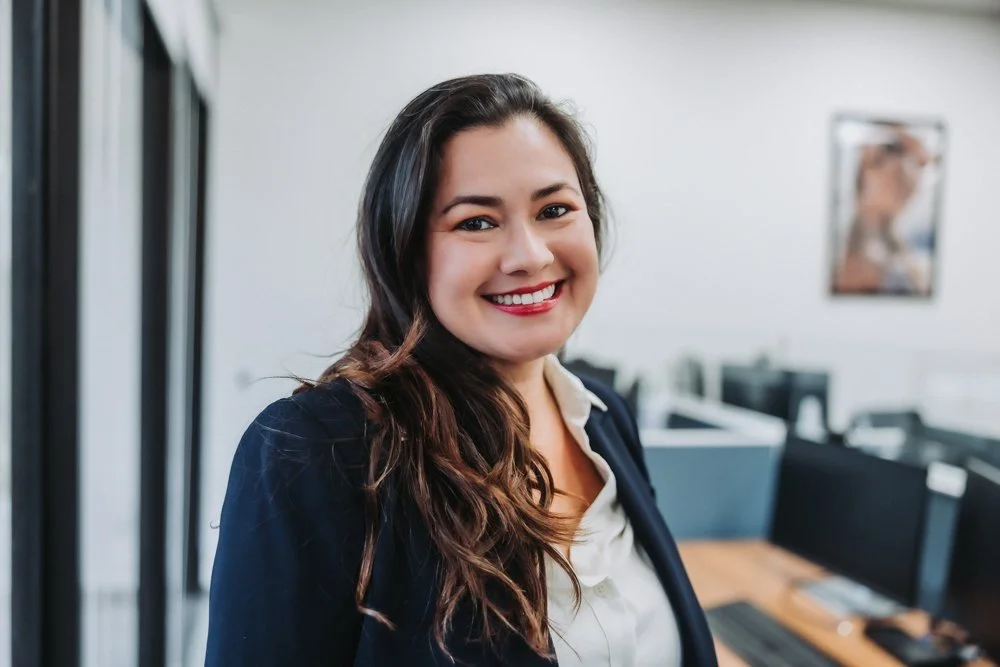 A professional woman with long, dark hair and a bright smile, wearing a navy blazer and white blouse, in an office setting.