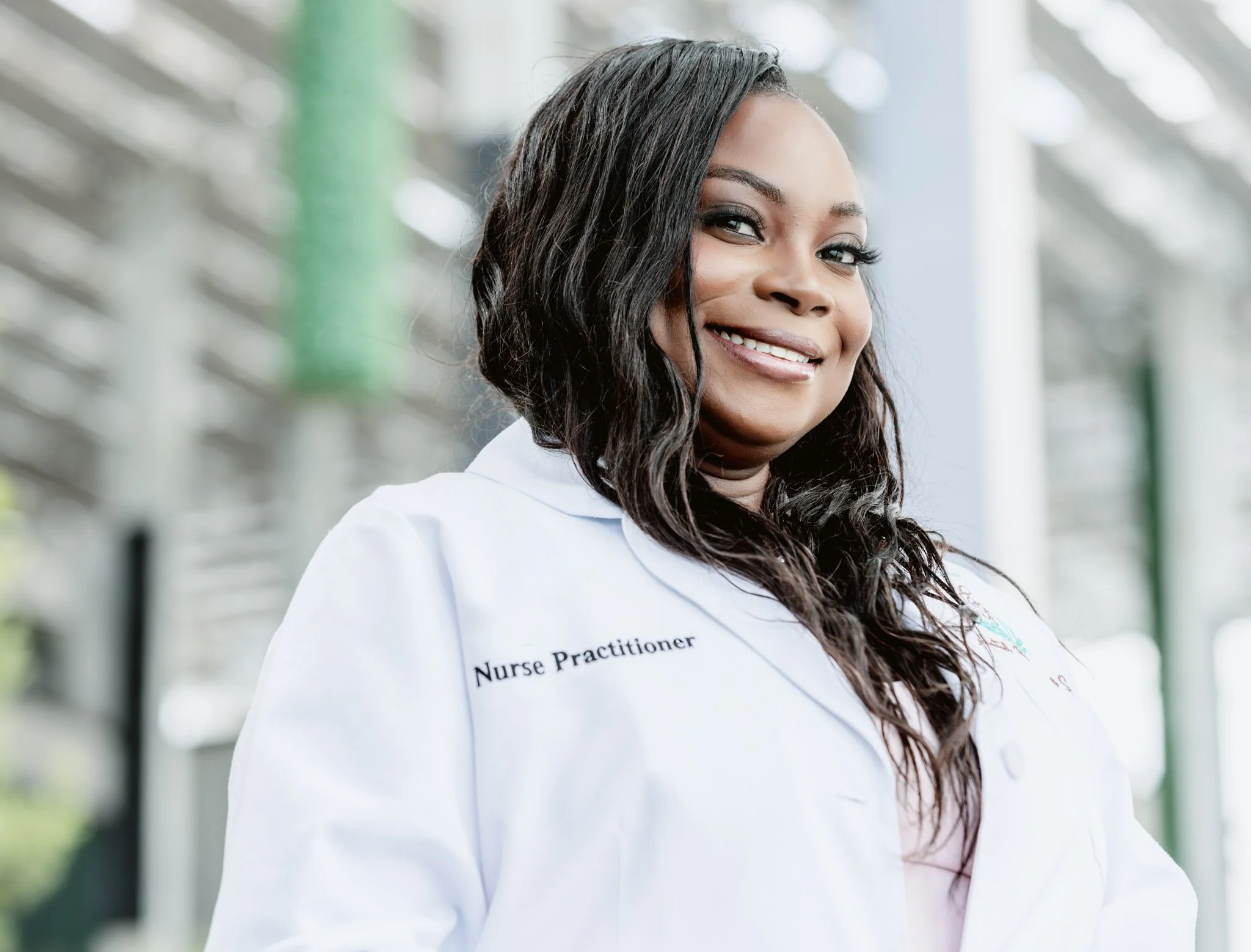A smiling woman with long, wavy black hair wearing a white healthcare uniform with the text "Nurse Practitioner" embroidered on it, standing in a modern building with glass and steel architecture.