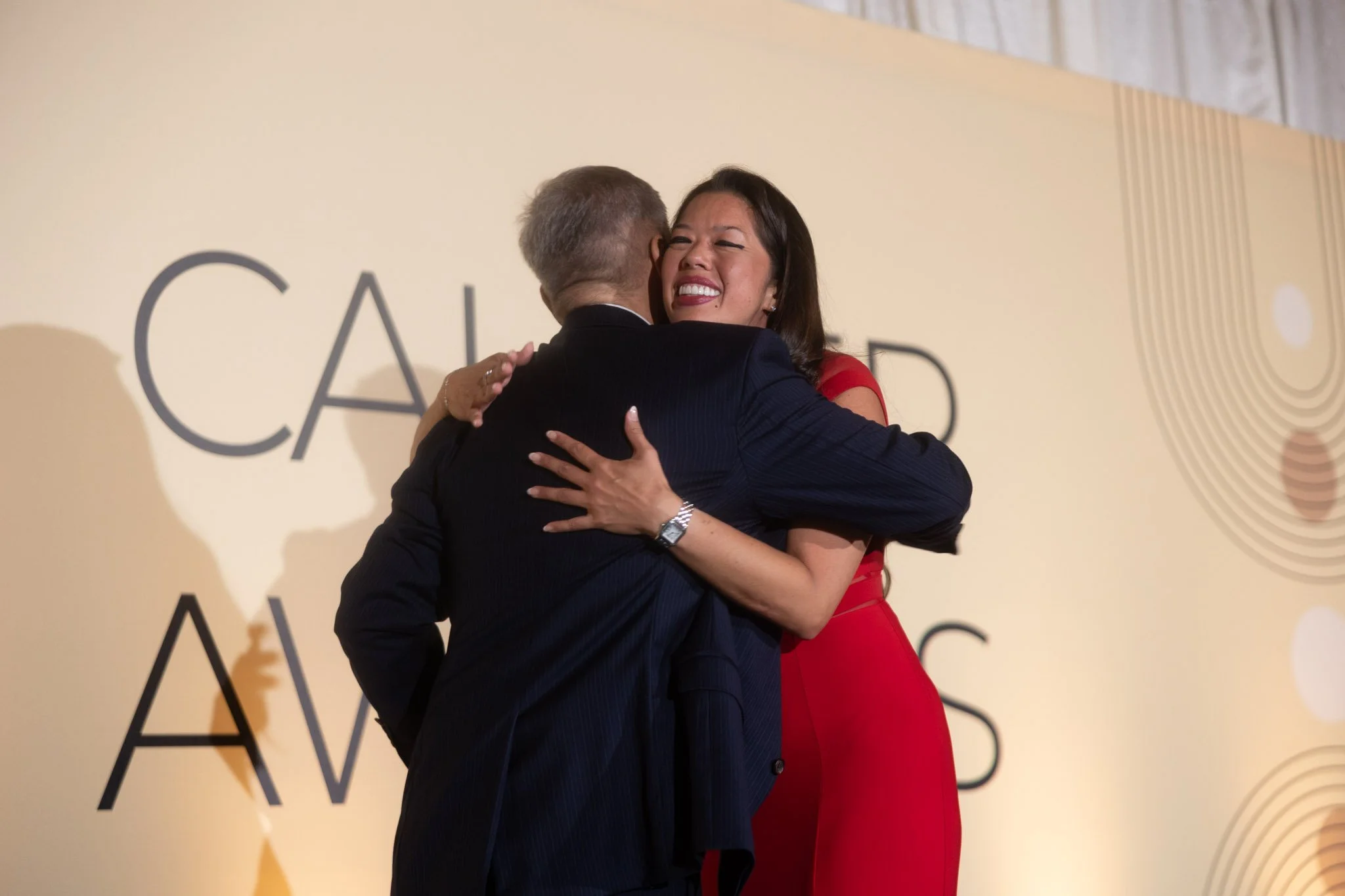 A woman in a red dress is hugging an older man in a tuxedo at an event with a beige backdrop displaying black text.