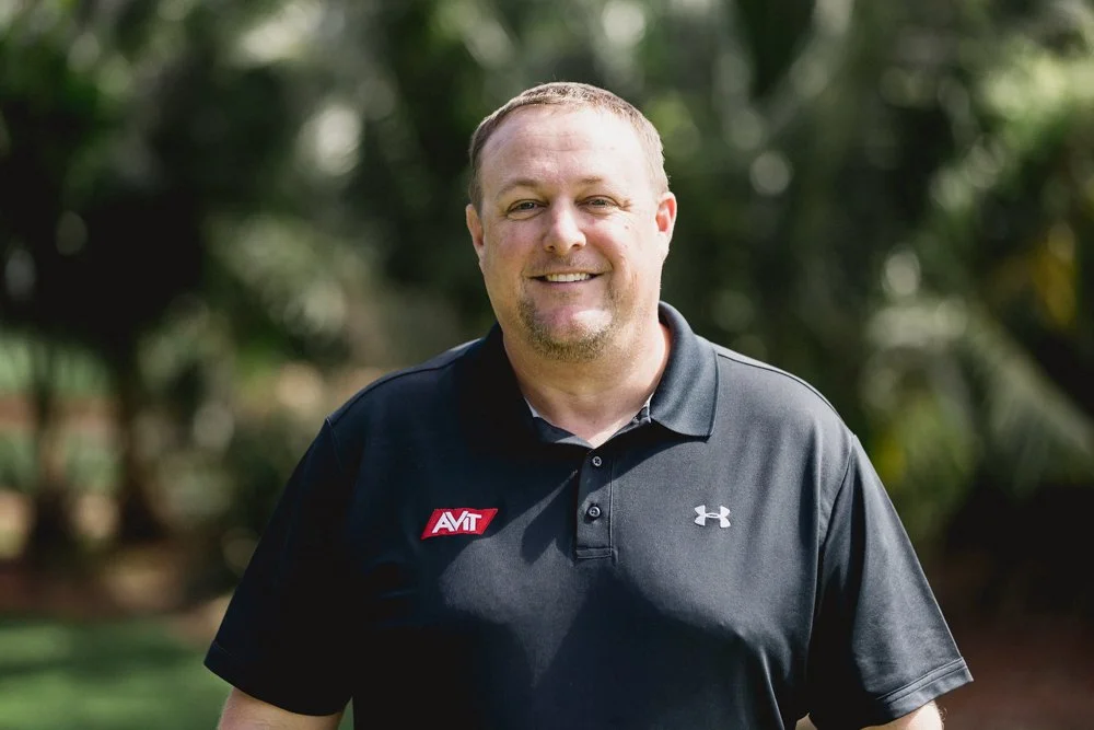 A smiling man standing outdoors with green trees in the background, wearing a black polo shirt with an AVT logo and an Under Armour logo.