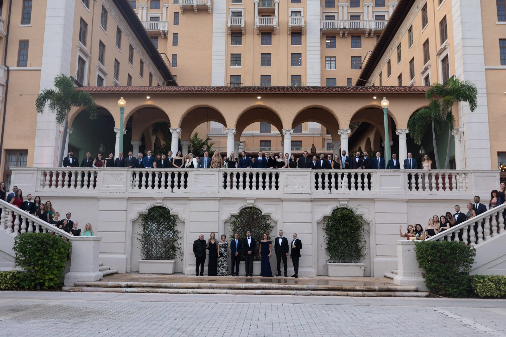 A large group of formally dressed people standing on an outdoor staircase and terrace in front of a grand, beige-colored building with arched columns and multiple stories.