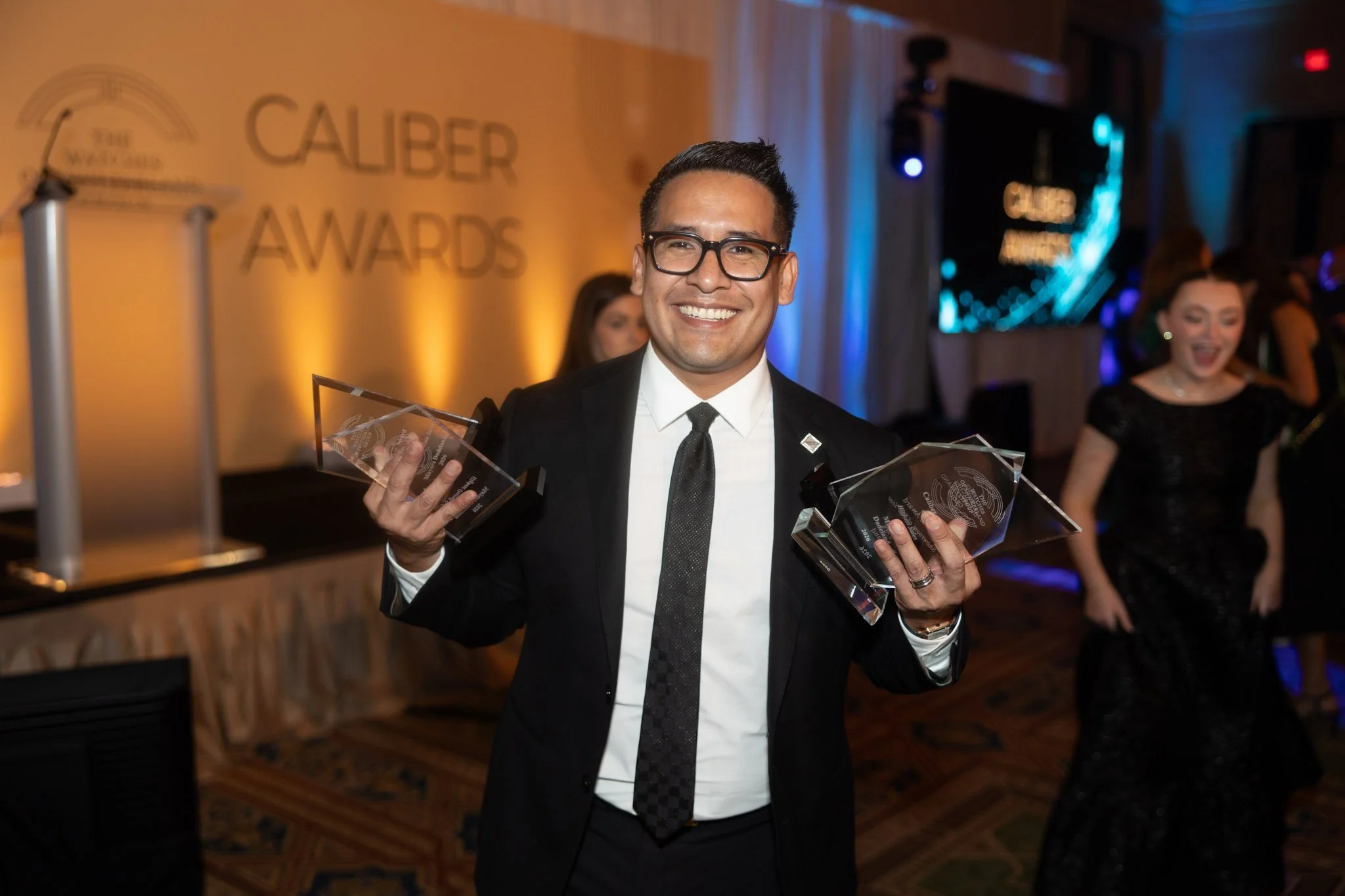 A man dressed in formal attire with glasses, smiling and holding multiple awards at an awards ceremony. In the background, there's a banner that reads 'The Caliber Awards' and other guests are celebrating.